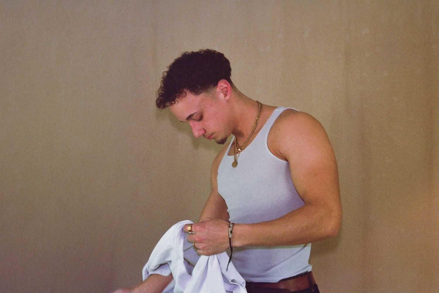 Muscular young man in a white tank top examining clothing while wearing a necklace and bracelet indoors.