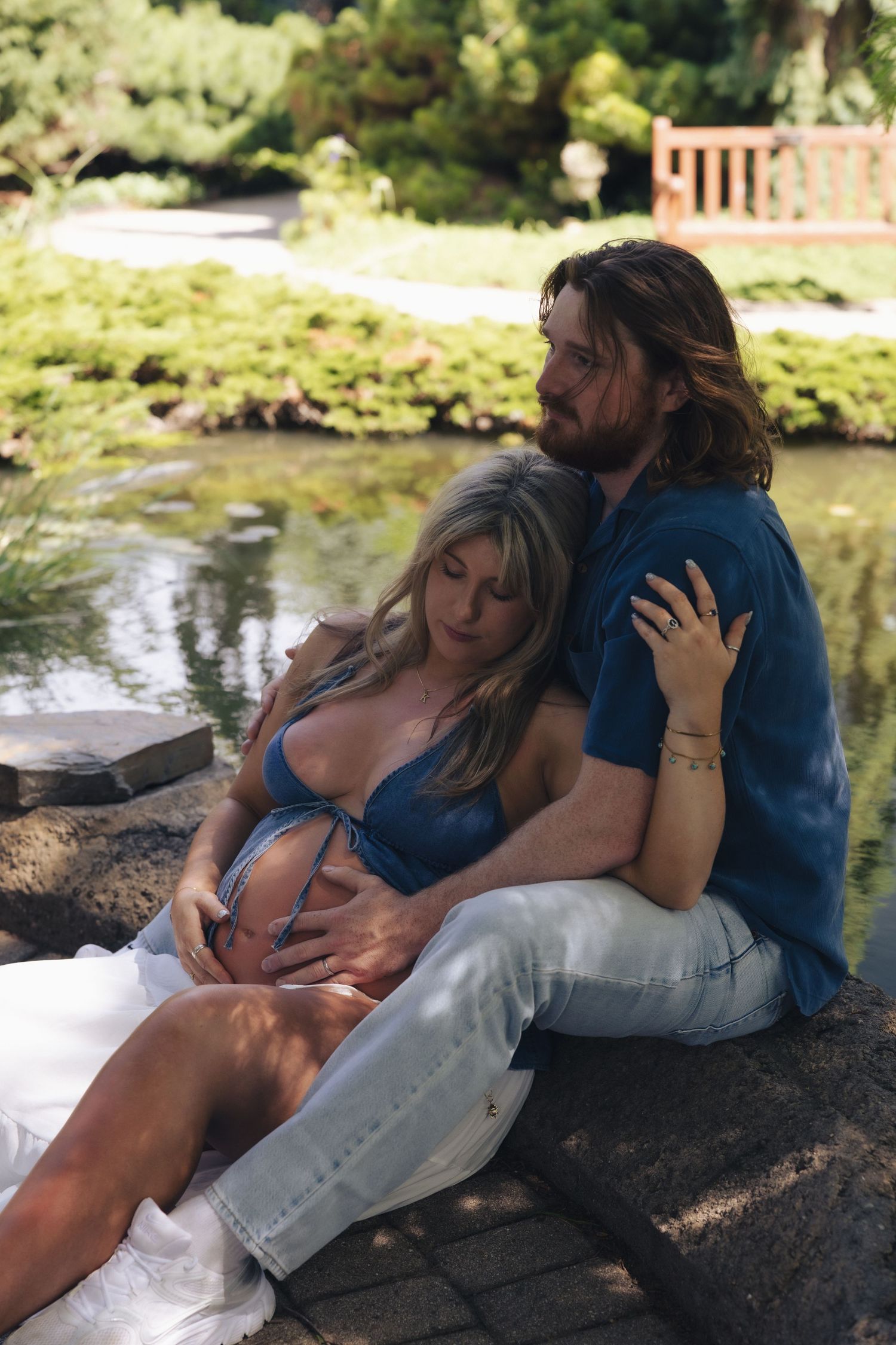 Couple sitting by a garden pond, man with long hair embracing pregnant woman in blue top outdoors.