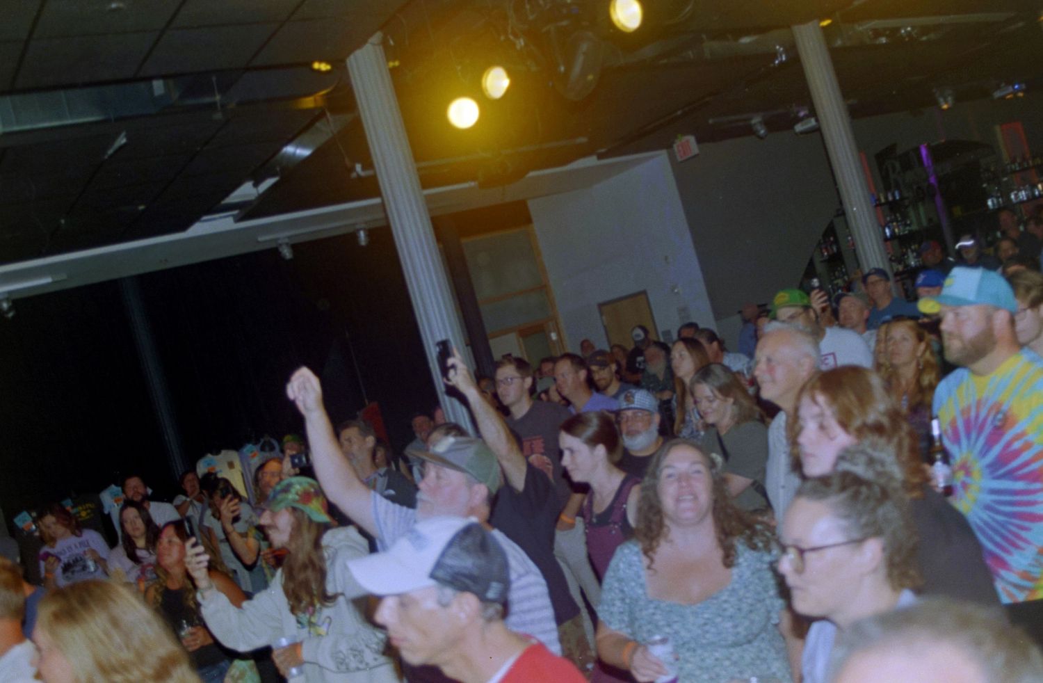Crowded indoor concert venue with audience members dancing and raising hands under stage lights.