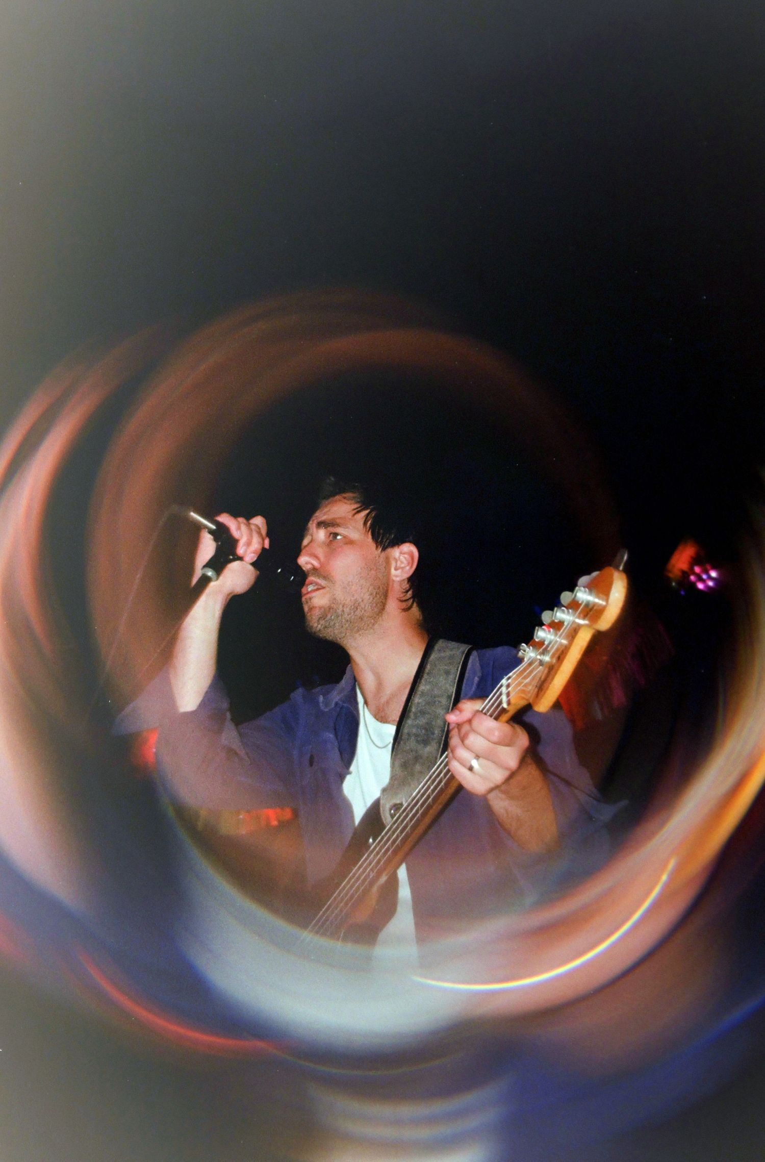 Male vocalist singing into microphone while playing bass guitar, captured with swirling light painting effect at live concert.