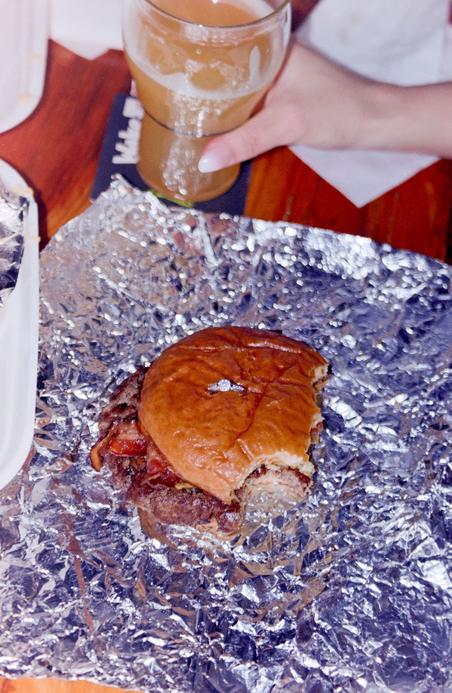 A juicy BBQ sandwich on a brioche bun served on crinkled aluminum foil with a glass of beer in the background.