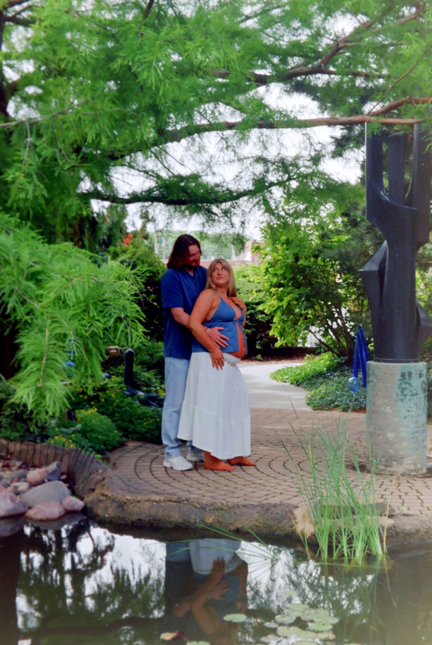 Pregnant couple posing by a garden pond surrounded by lush greenery and a stone pathway.