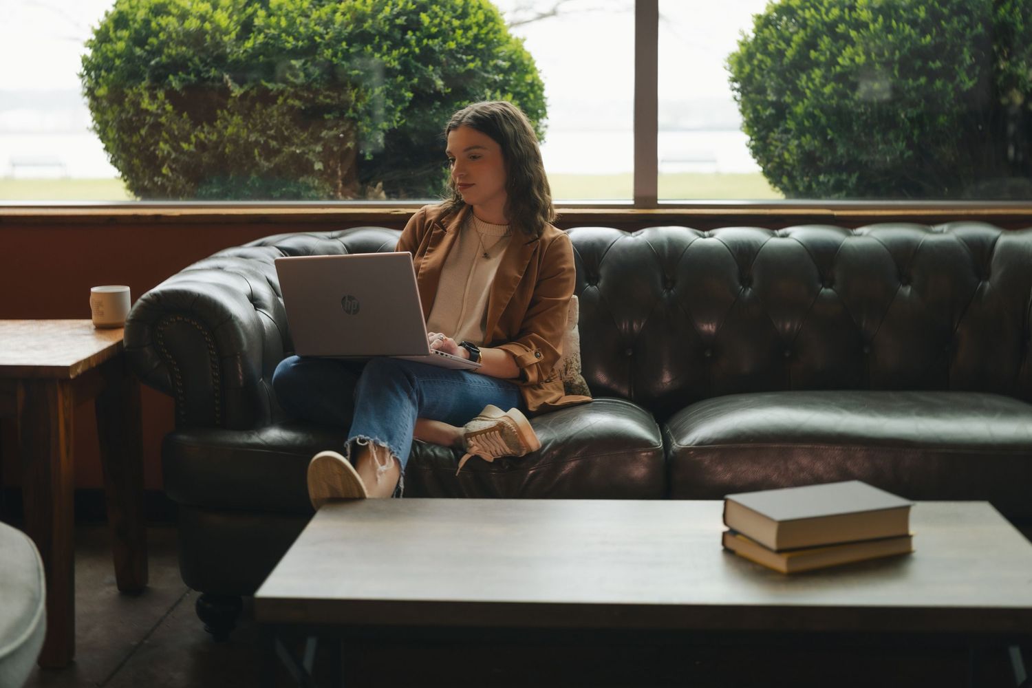 Woman sitting on leather sofa using laptop computer in cozy cafe with large windows and books on table.