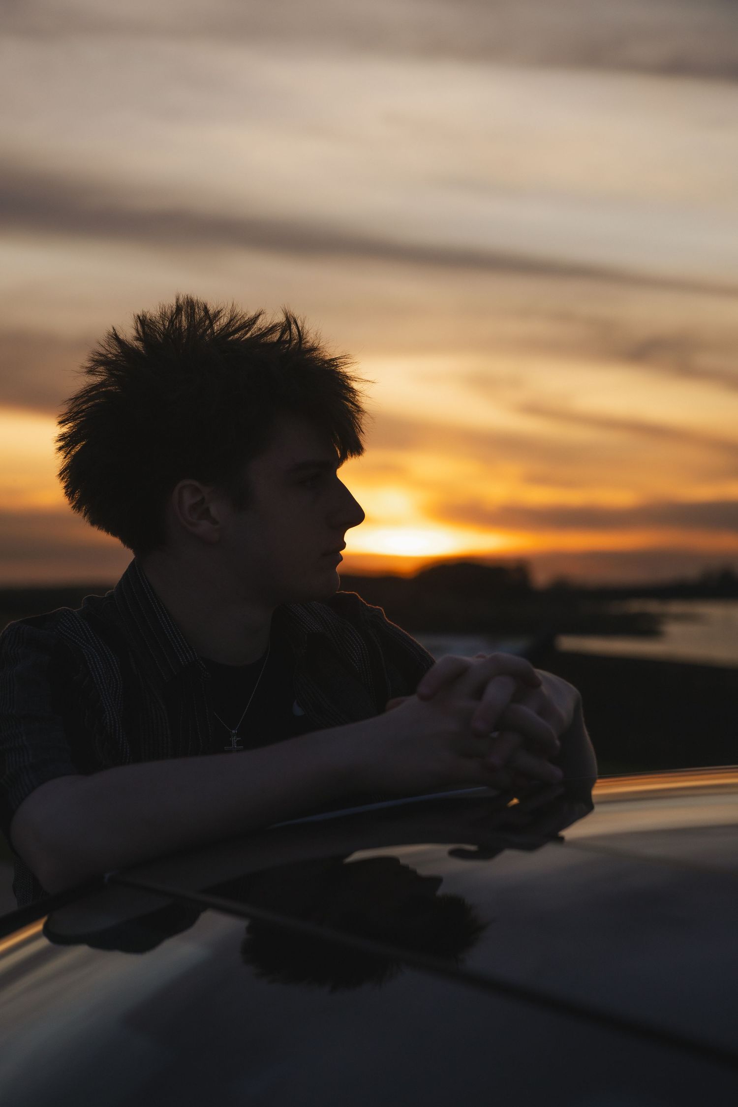 Silhouette of a young man resting his hands on a car roof, gazing at a dramatic golden sunset sky.