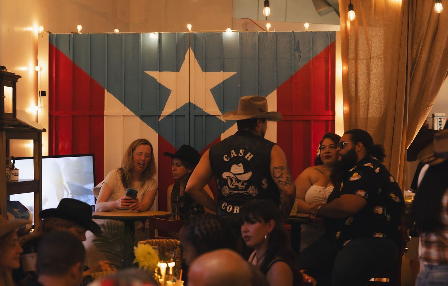 People socializing in a bar with a large Puerto Rican flag mural, string lights, and a man in a cowboy hat.