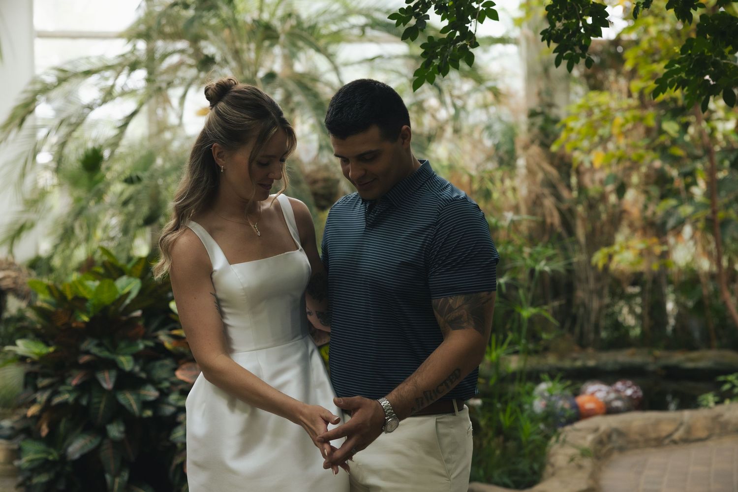 Couple holding hands in a lush greenhouse, woman in white dress and man in blue shirt with tattoos.