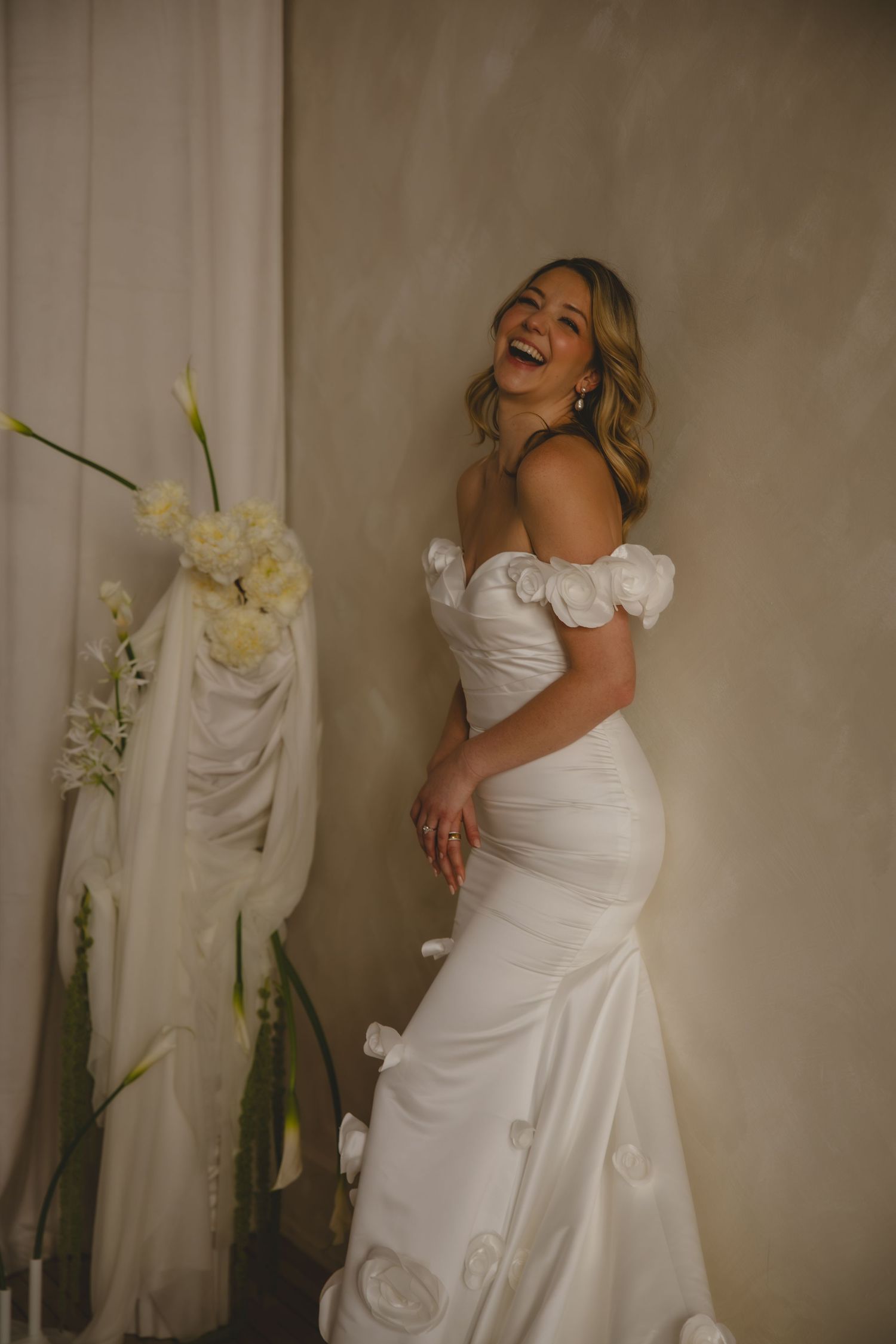 A joyful bride in an elegant off-shoulder white wedding gown laughs while standing beside floral decorations.