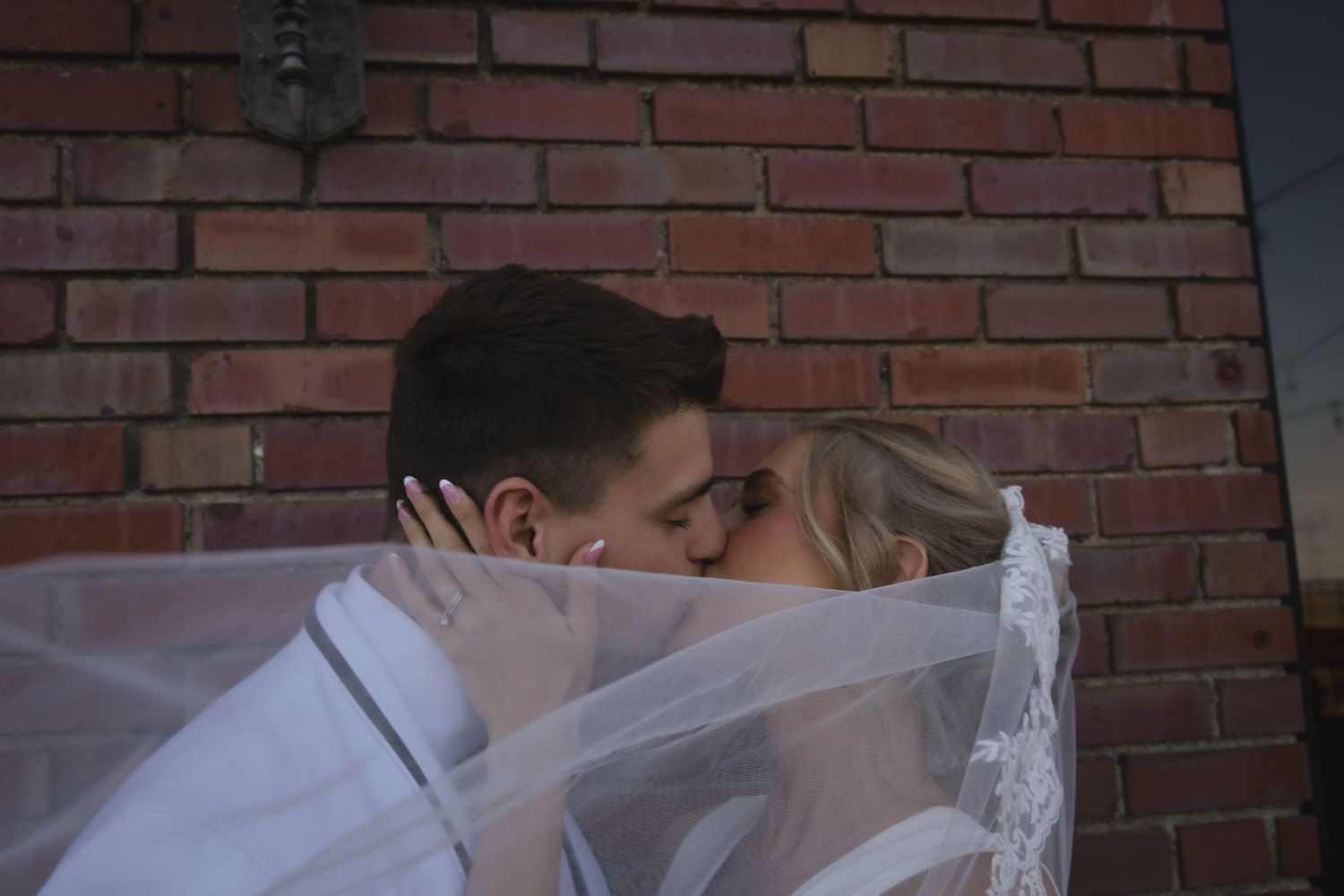A romantic wedding couple sharing a kiss under flowing white veil against rustic brick wall background.