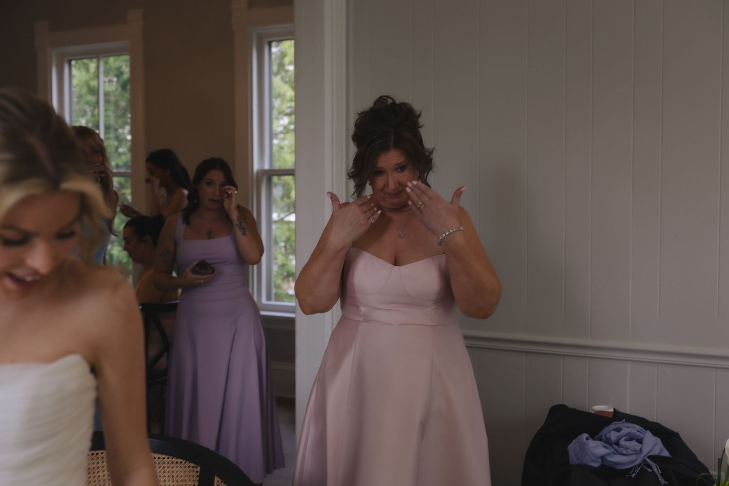 Emotional bridesmaid in pink dress wiping tears while bridesmaids in lavender dresses stand in background during wedding preparations.