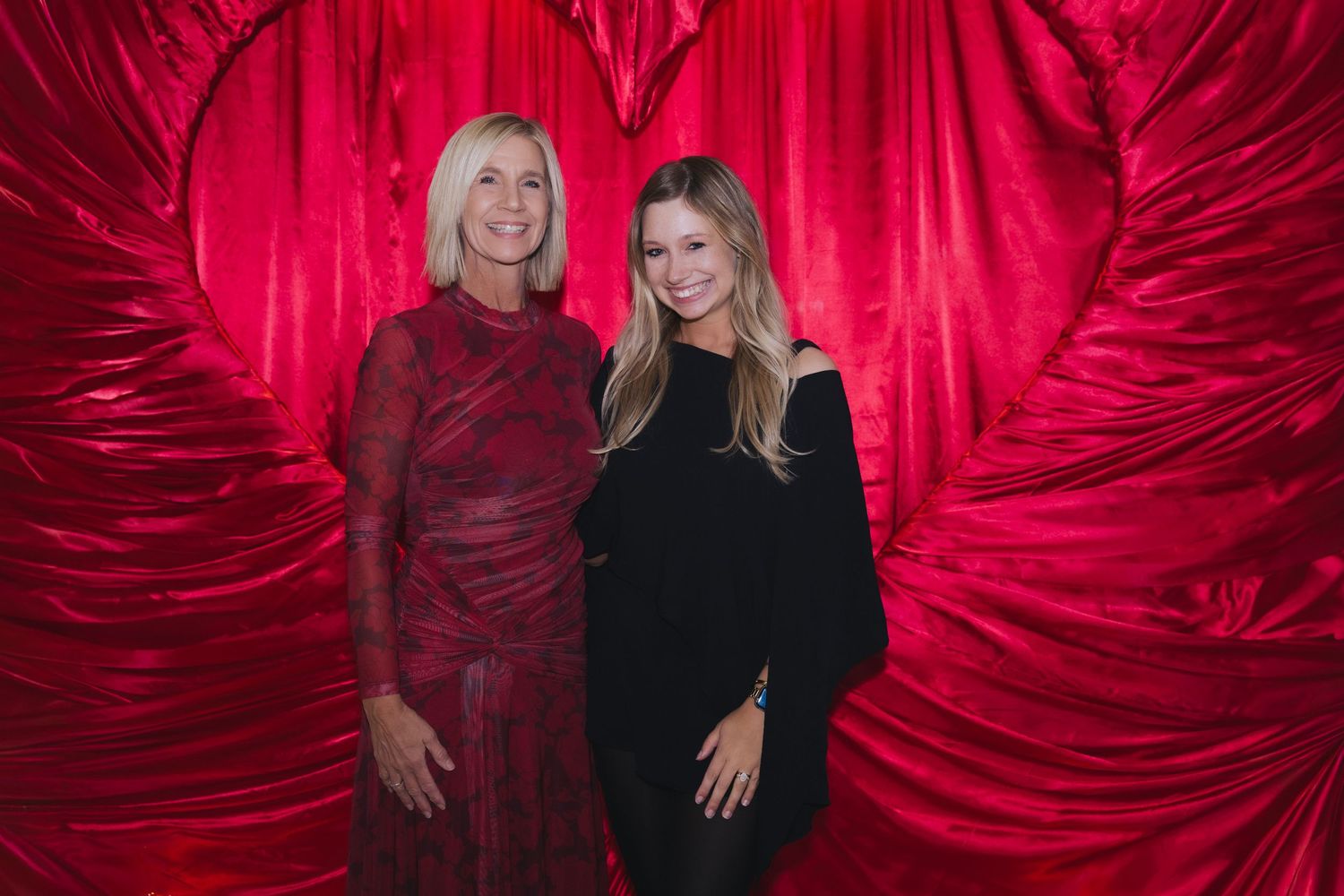 Two people pose together against a dramatic red velvet curtain backdrop in elegant evening attire.