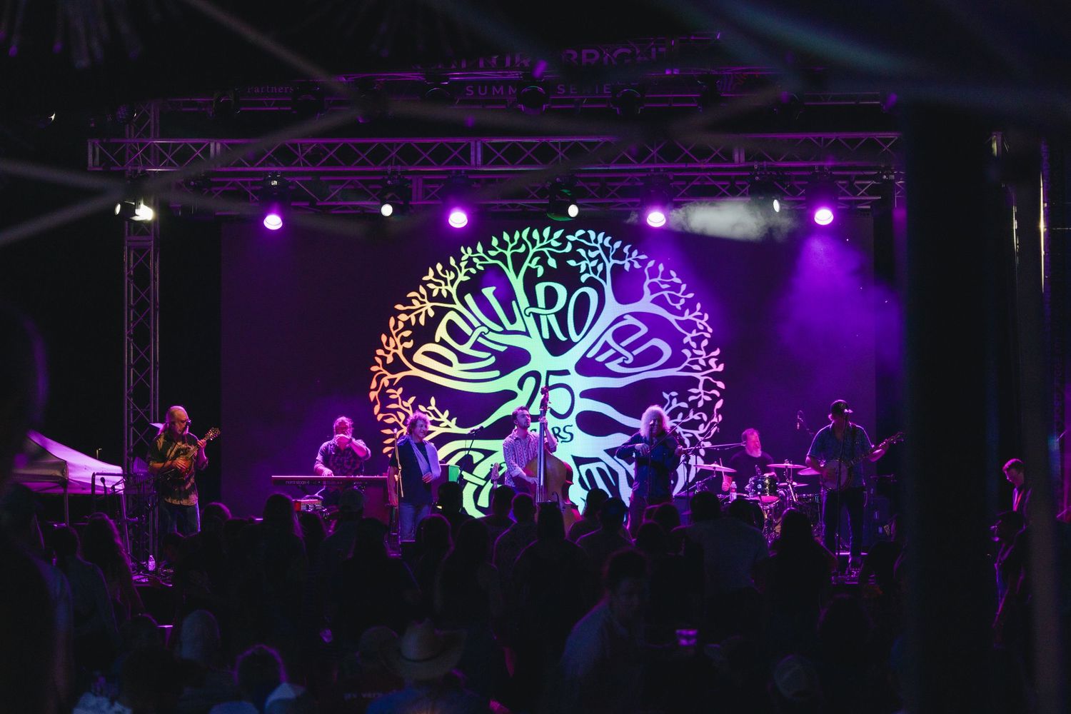 Live band performs on a vibrant purple-lit stage with glowing tree logo backdrop before a silhouetted crowd.