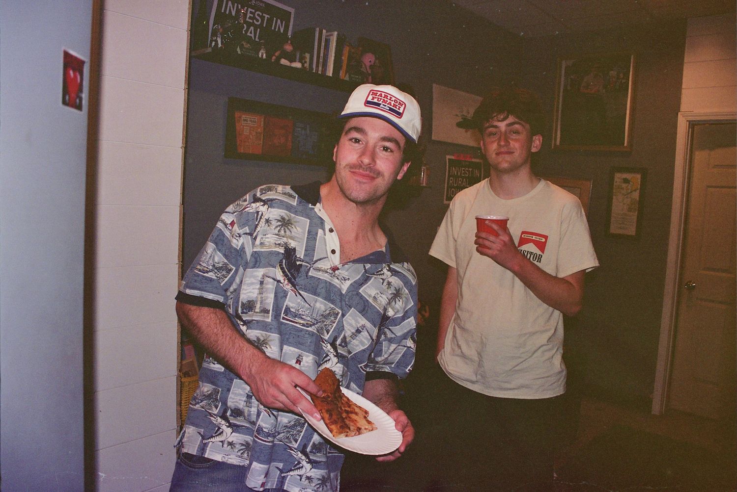 Two young men at a casual party, one wearing a Hawaiian shirt and cap holding a plate of food, the other in a white tee.