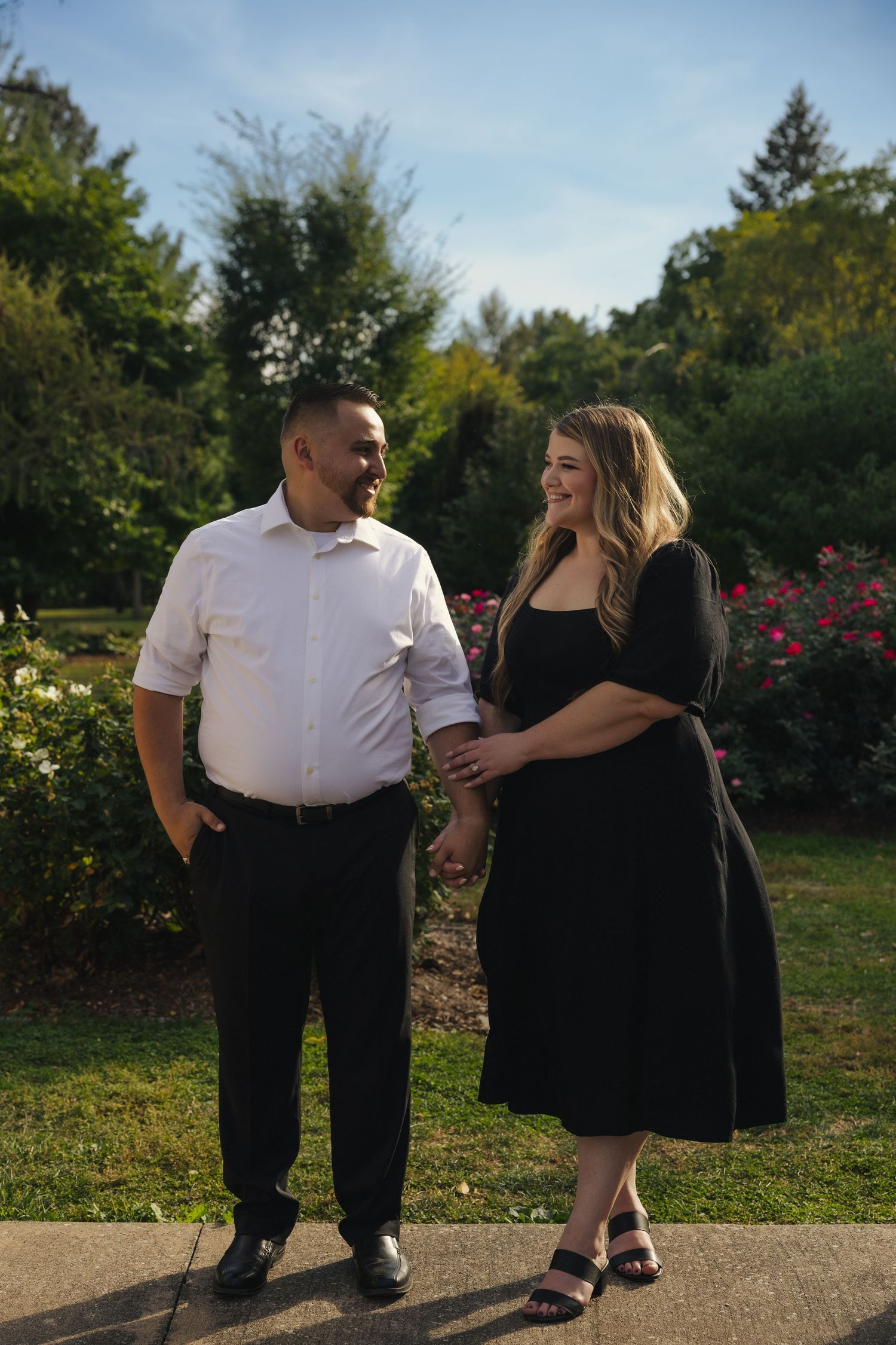 Couple holding hands and smiling in a garden with roses and greenery, man in white shirt, woman in black dress.
