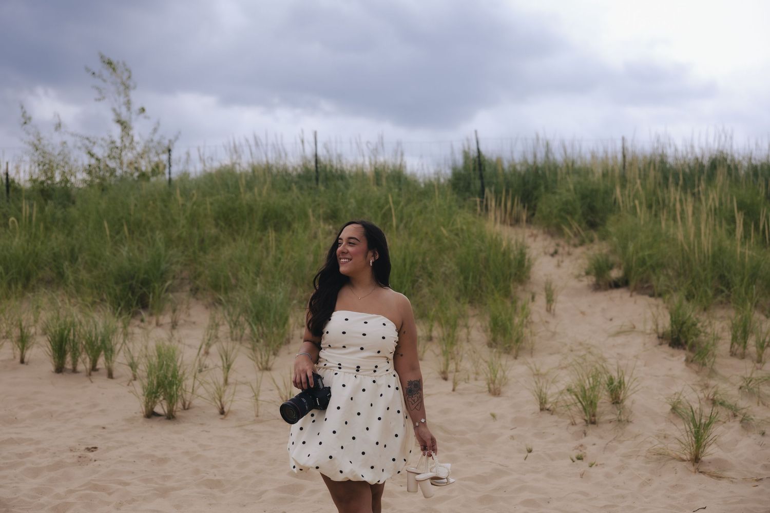 Woman in white polka dot dress holding camera walks on sandy beach dunes with tall grass under cloudy sky.