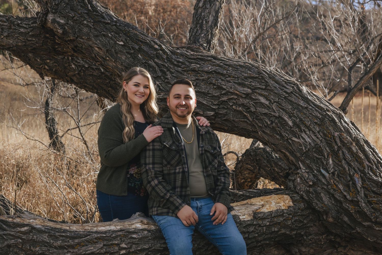 Smiling couple sitting on a large tree trunk in a rustic outdoor setting with dry grass and trees in the background.