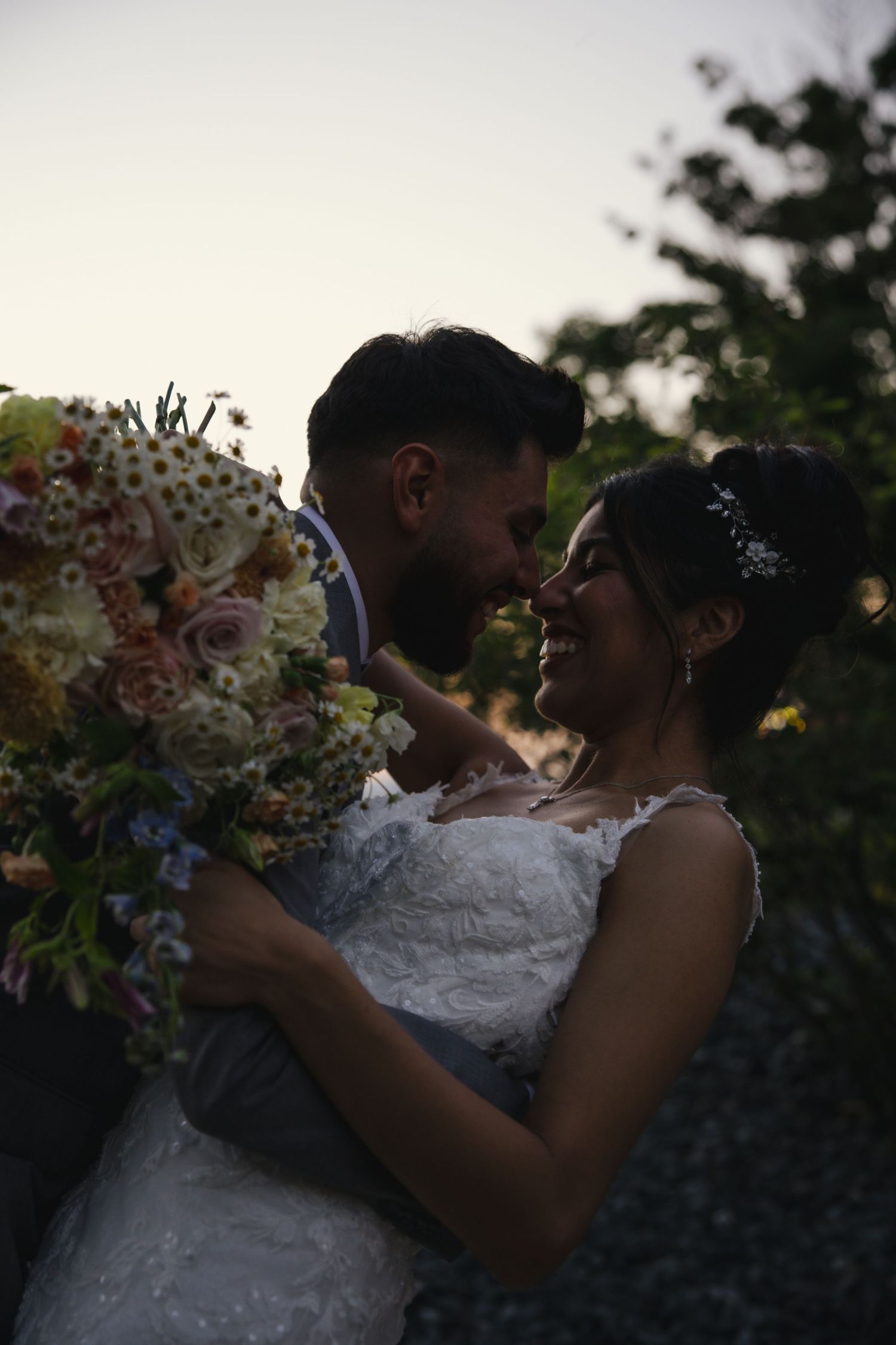 A couple in wedding attire shares tender kisses at sunset with a bouquet of flowers visible.