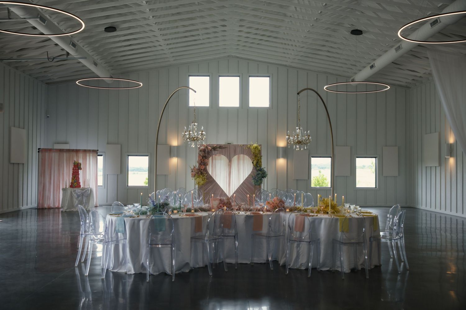 Elegant wedding reception venue with white tables, ghost chairs, and heart-shaped backdrop under circular chandeliers.