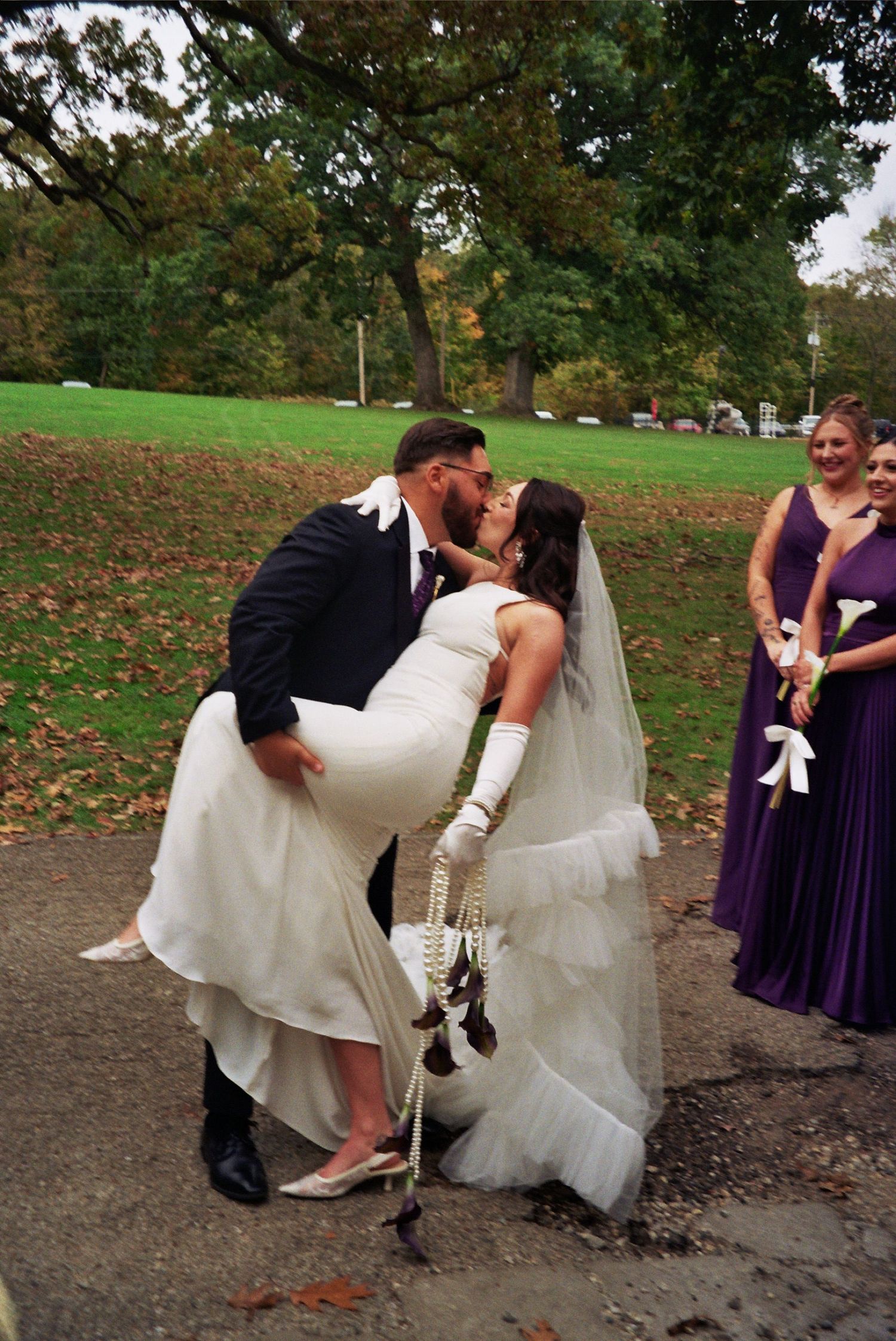 Bride and groom share a romantic dip kiss at outdoor wedding, with bridesmaids in purple dresses watching nearby.