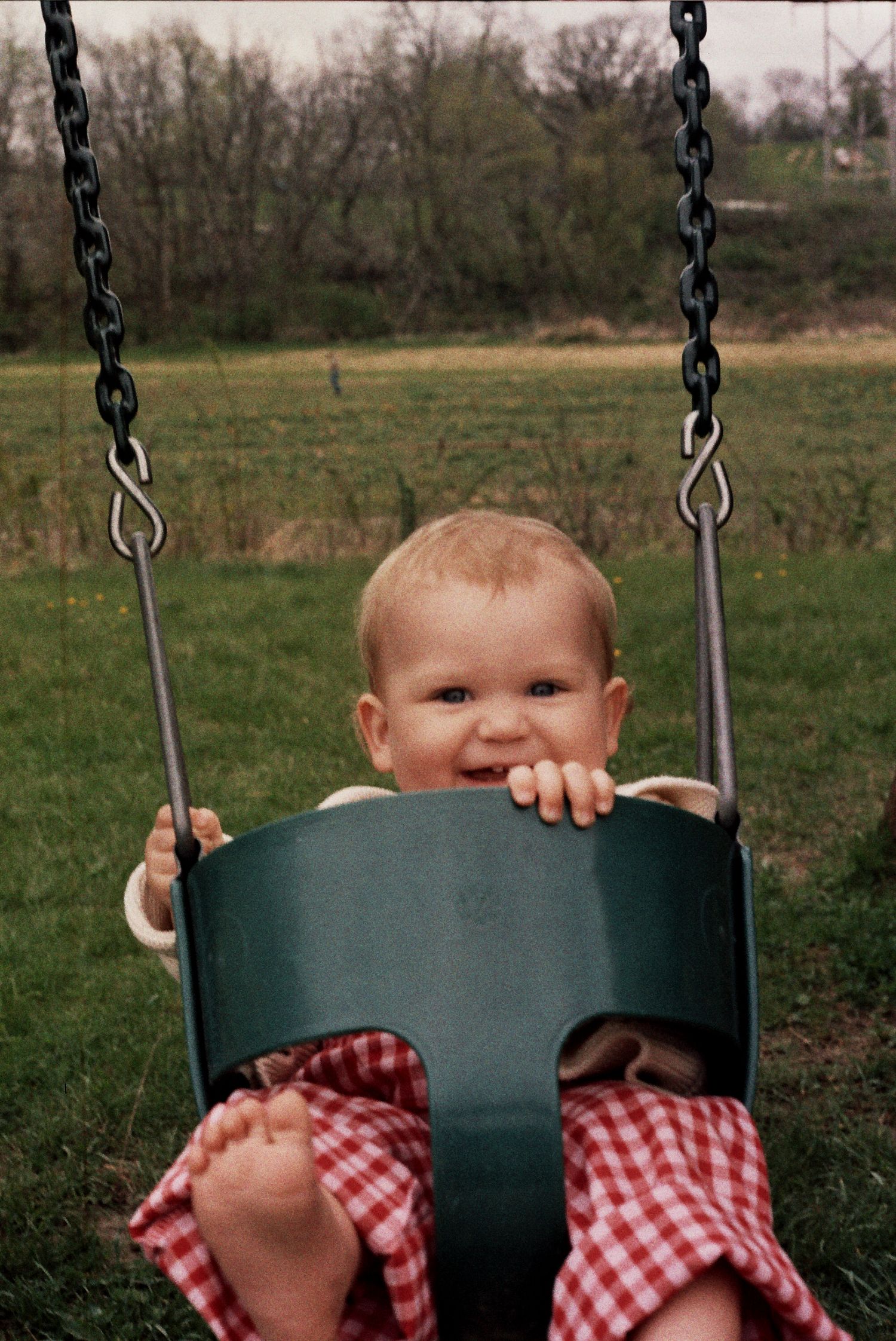 Happy baby sits in a green swing wearing red gingham with bare feet dangling in an outdoor setting.