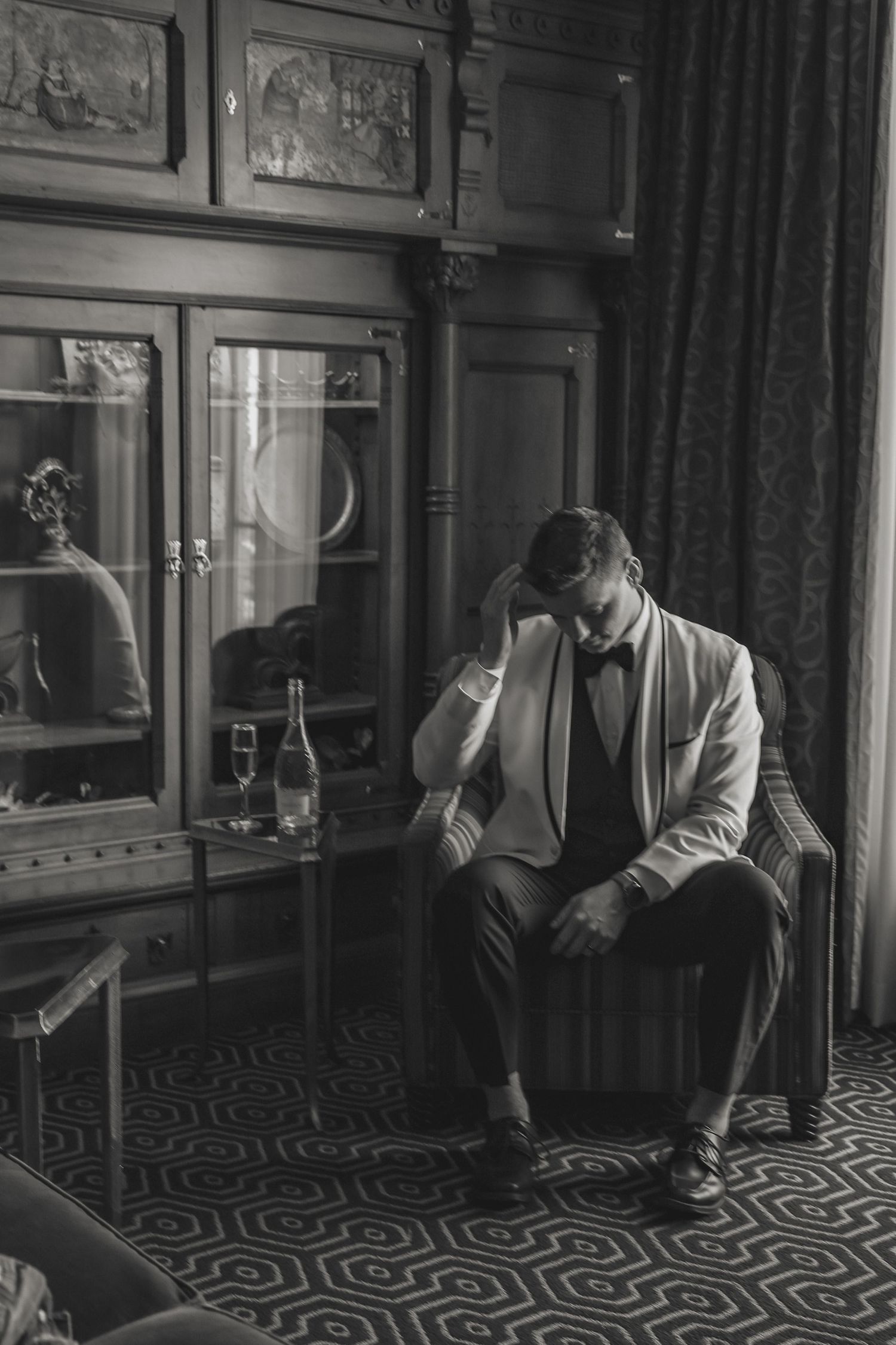 Black and white photo of person in light blazer sitting alone in vintage room with patterned carpet and antique furniture.