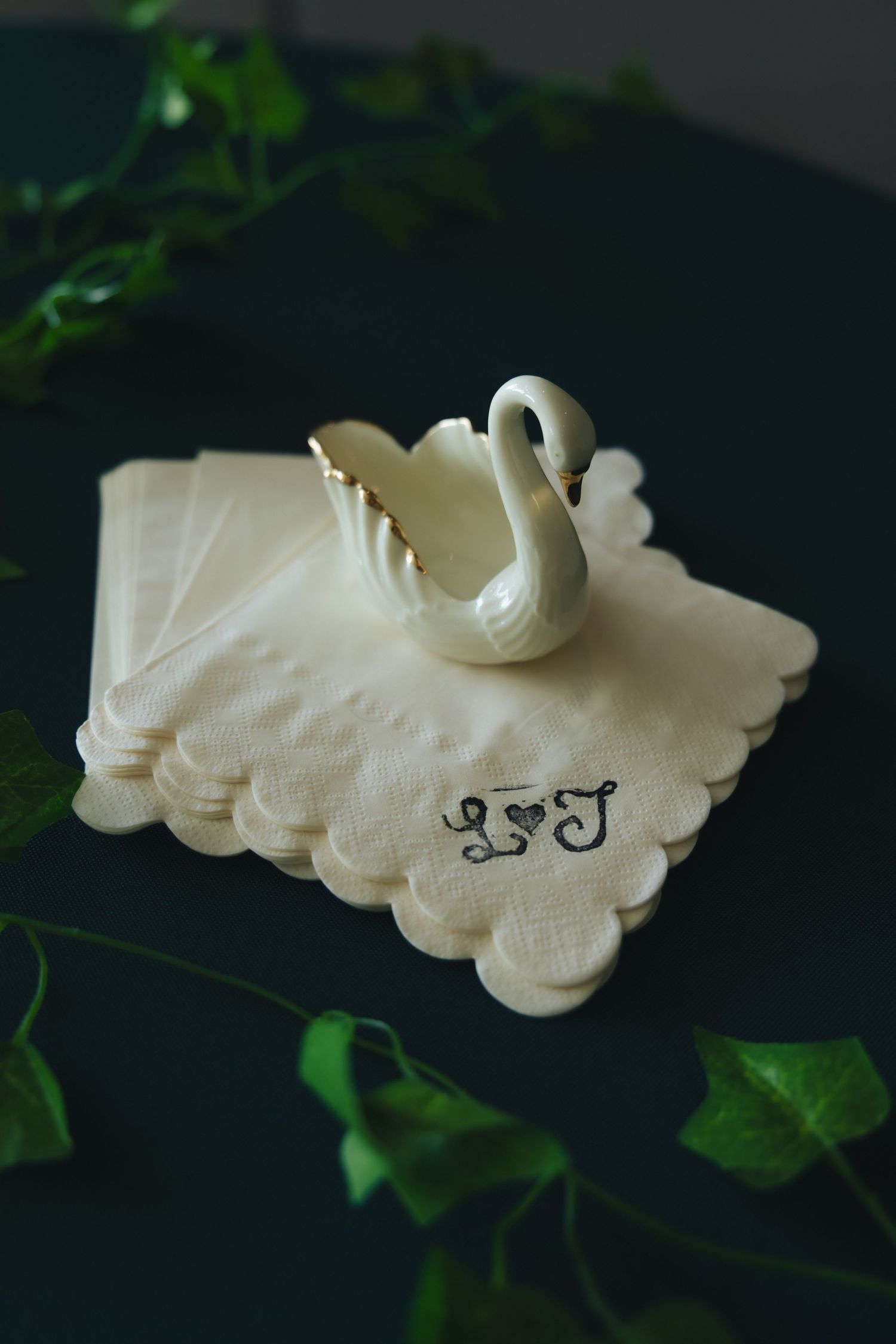 White ceramic swan figurine on monogrammed scalloped napkins with ivy on a dark green table.