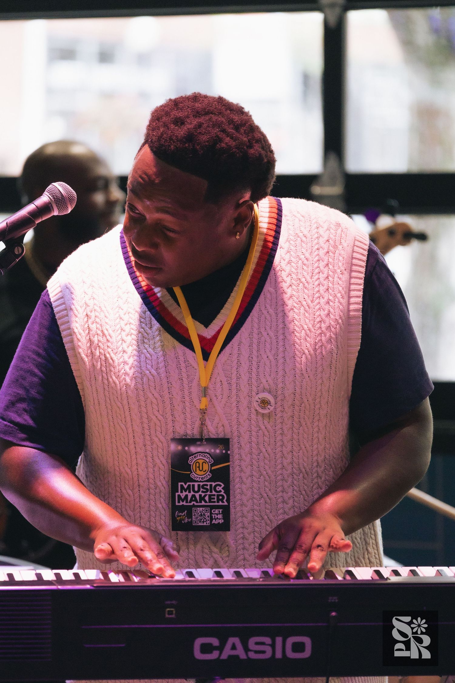 A musician plays a Casio keyboard wearing a white knit vest and Music Maker lanyard at an outdoor performance.