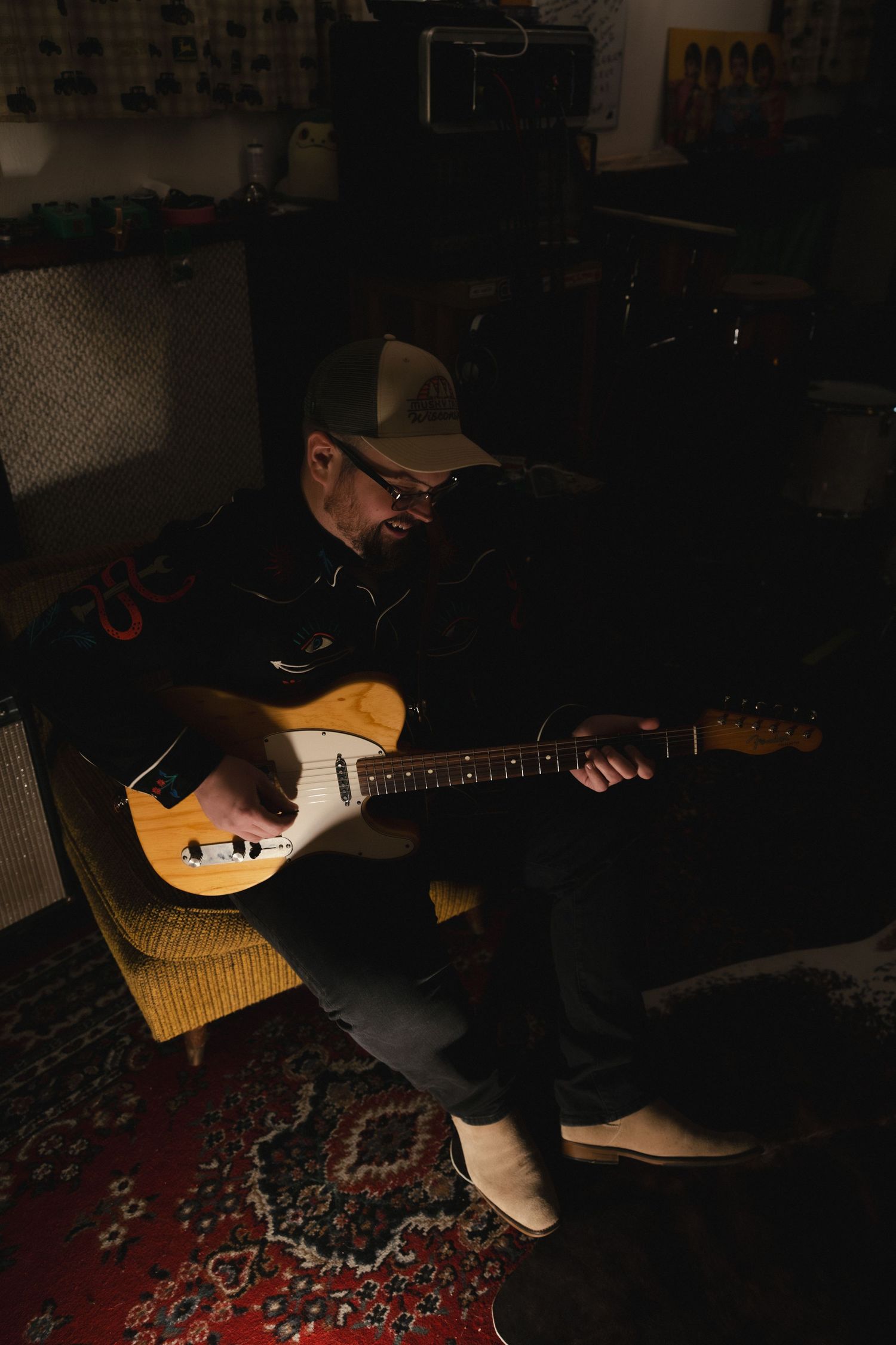 Man playing electric guitar in dimly lit room, wearing cap and glasses, seated in chair with amplifiers in background.