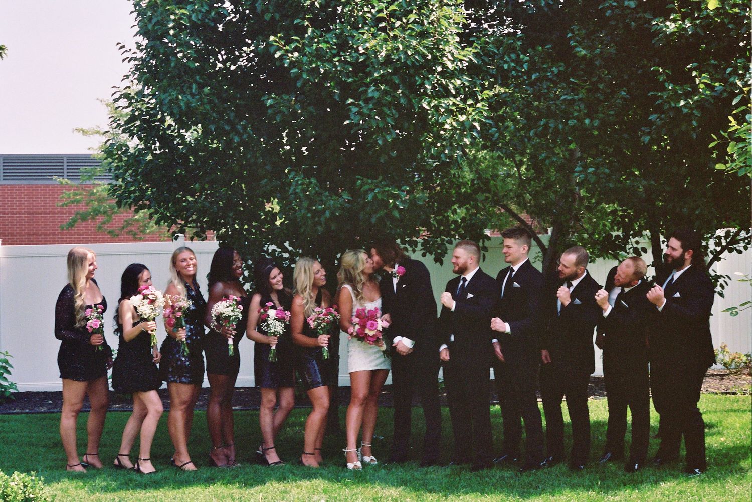 A wedding party in formal black attire poses outdoors under a green tree on a sunny day.