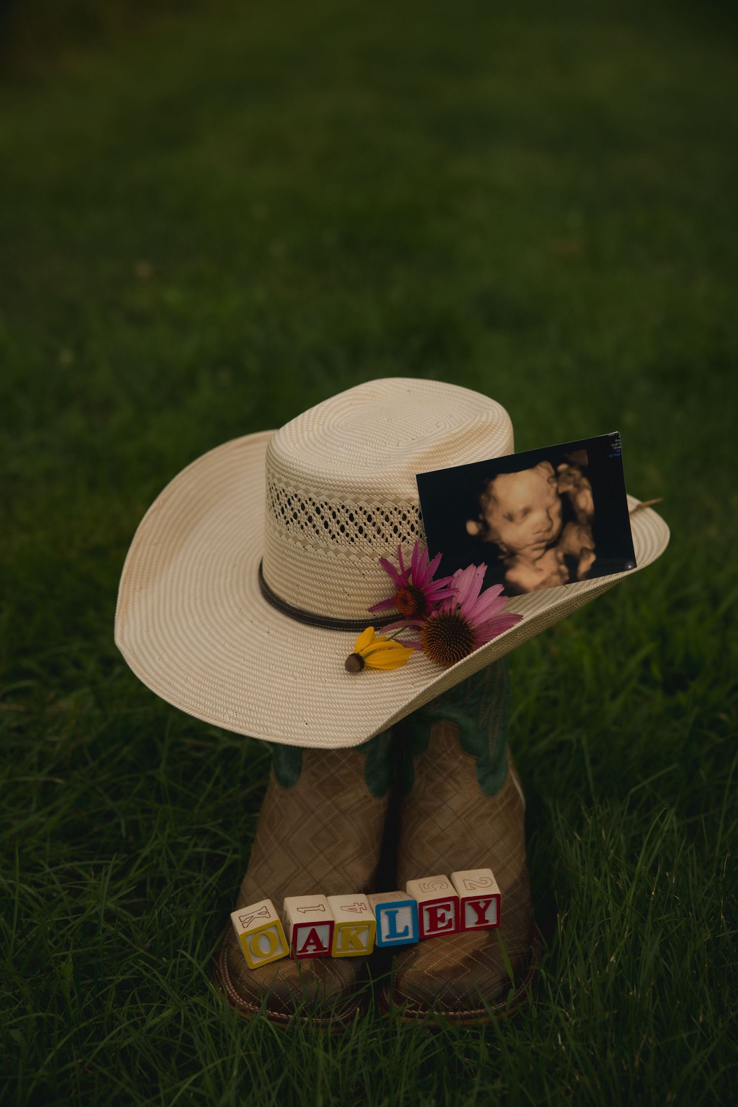A tan cowboy hat with flowers and photo rests atop brown boots with alphabet blocks spelling out a name on grass.