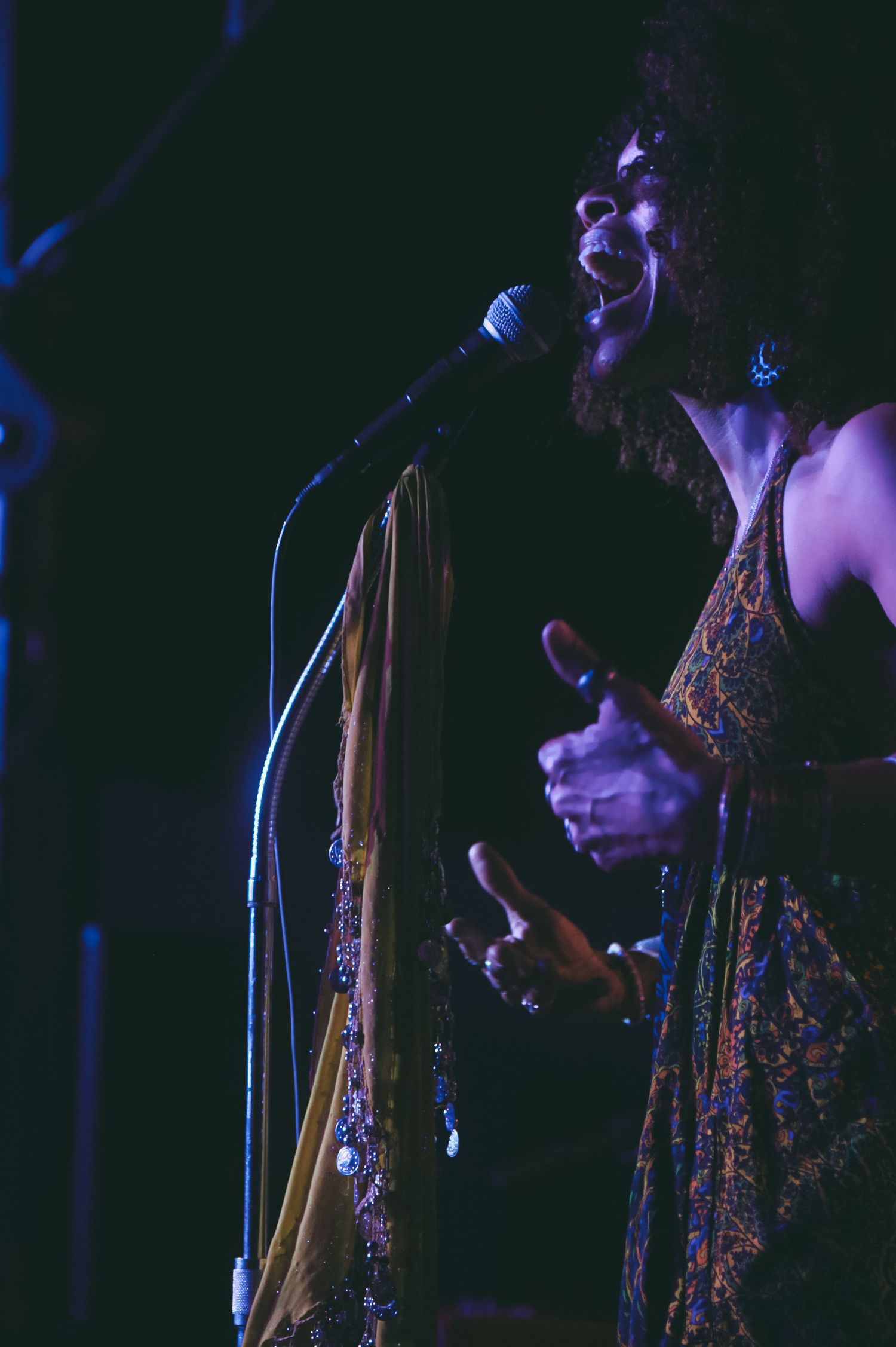 A performer in a patterned dress sings passionately into a microphone on a darkened stage.