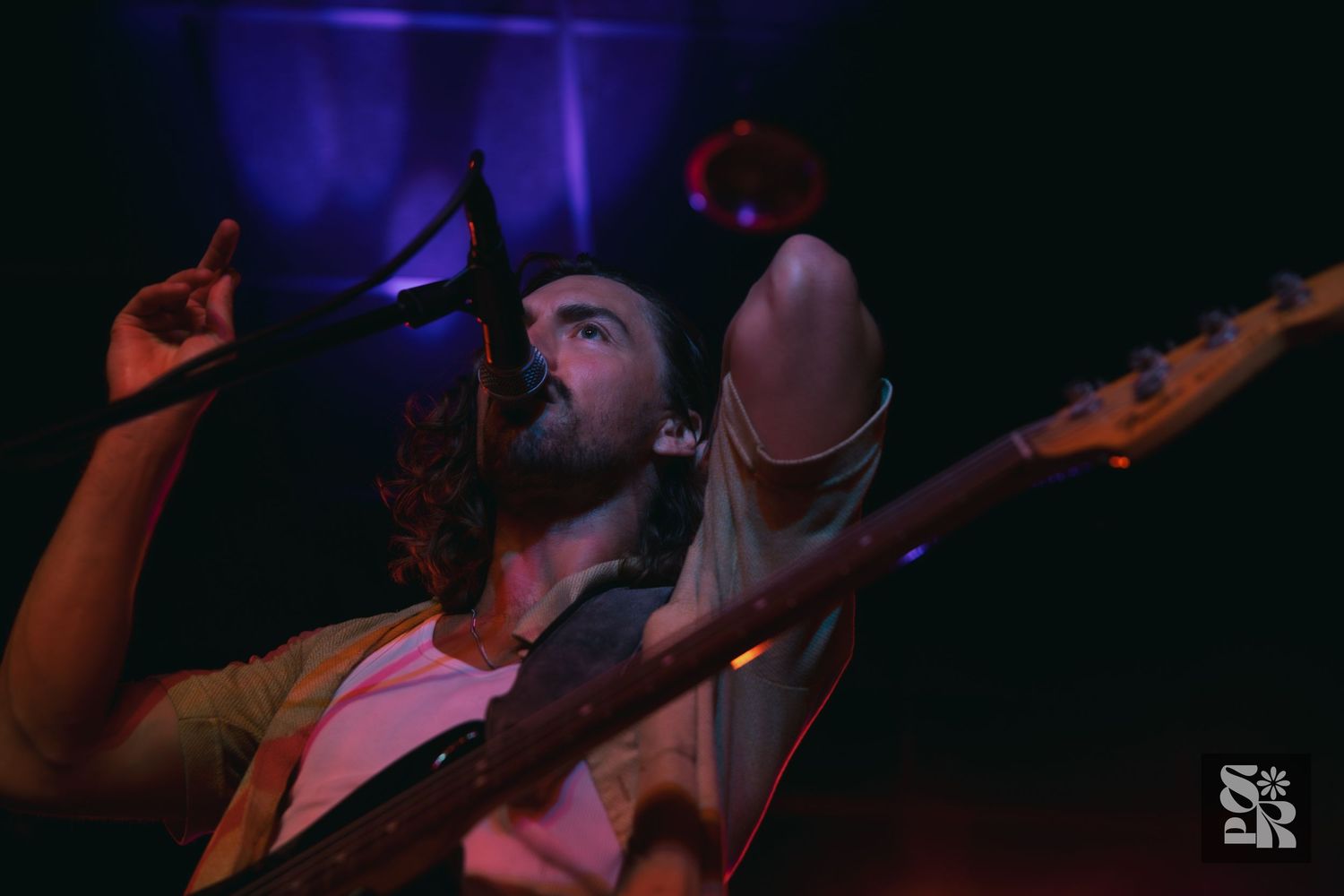 Concert performers playing guitars and singing on stage with dramatic purple and blue lighting effects.
