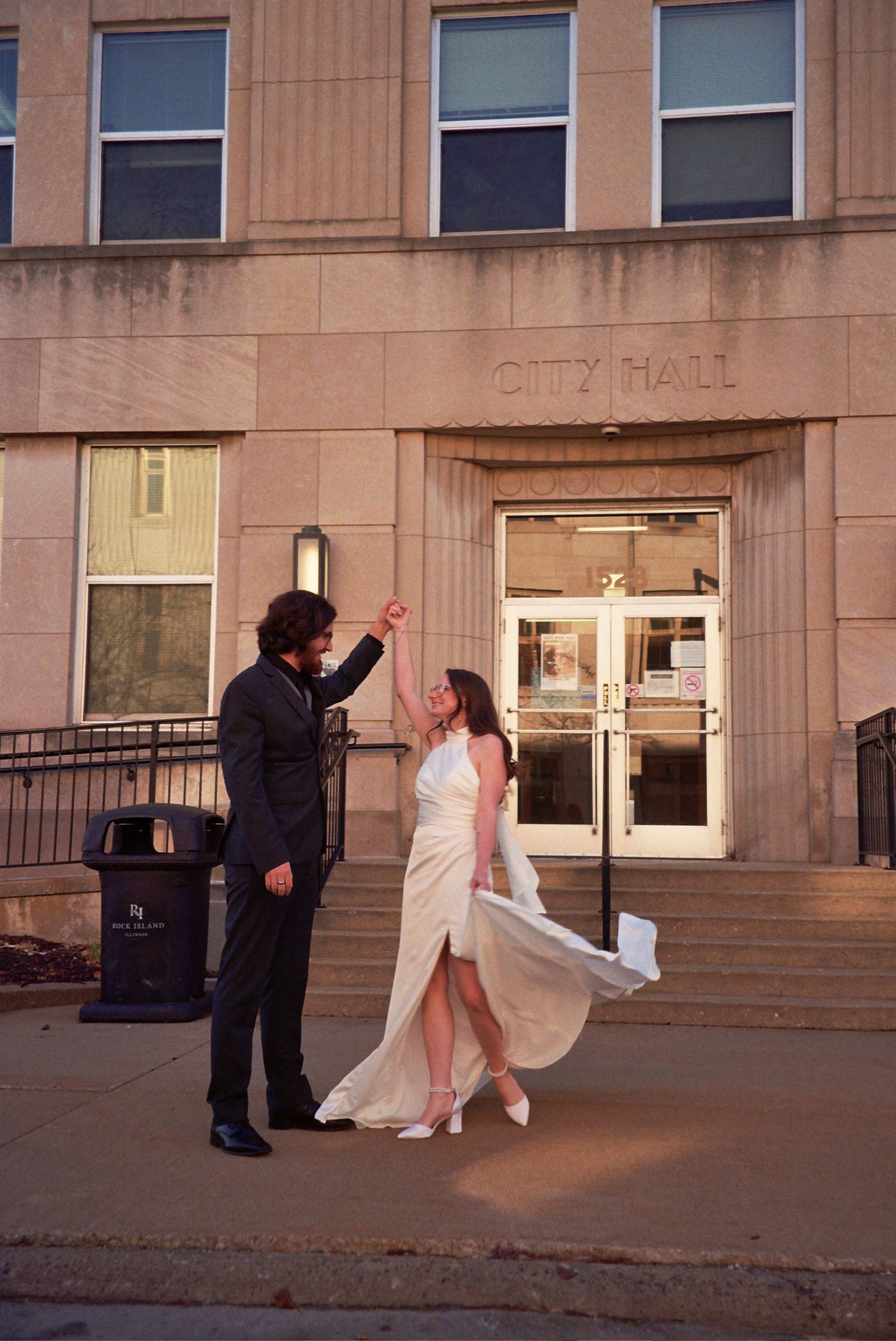 Newlywed couple dancing outside City Hall, bride in flowing white gown, groom in black suit, golden hour lighting.
