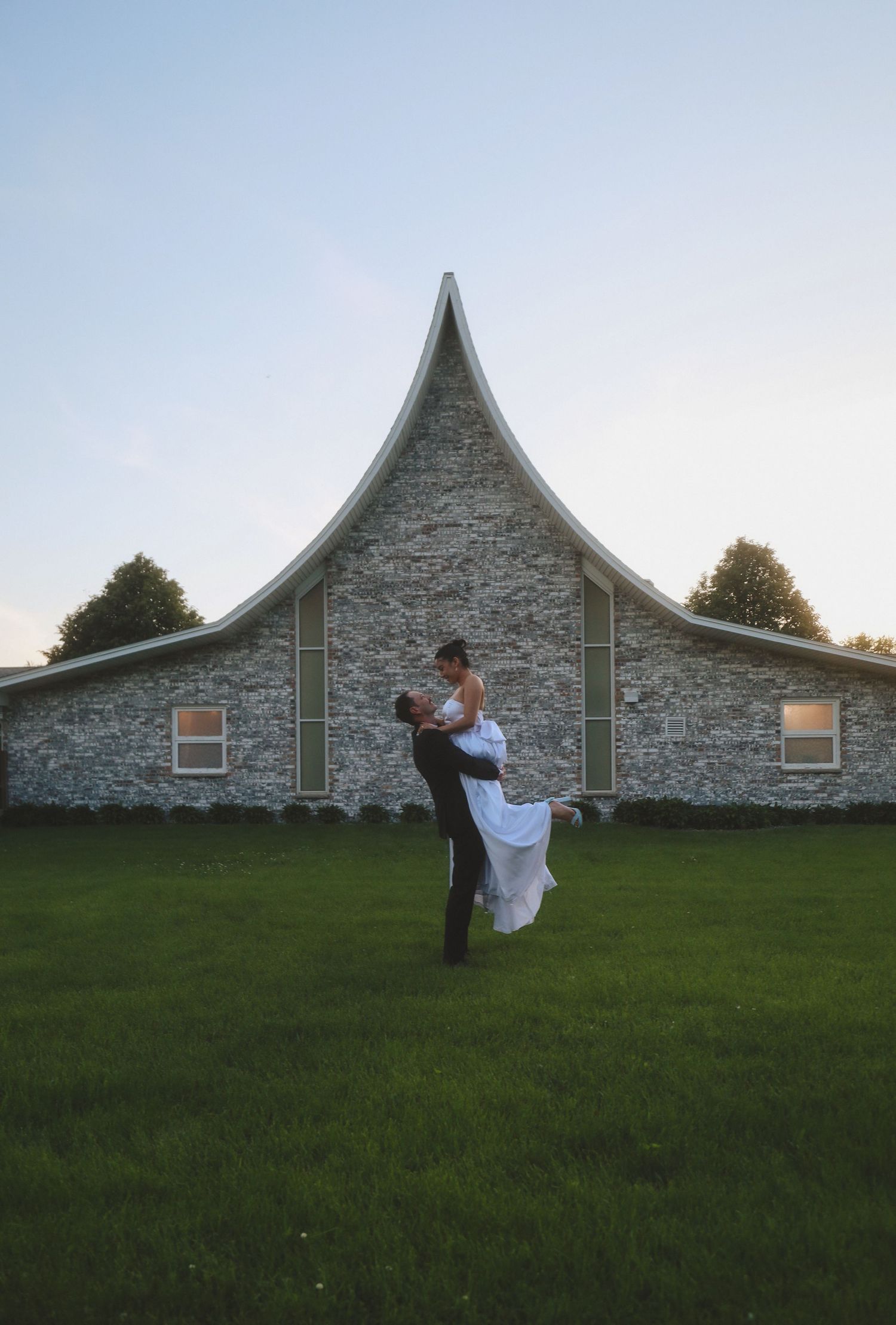 A couple shares a romantic moment outside a stone church with a dramatic peaked roof during twilight hours.