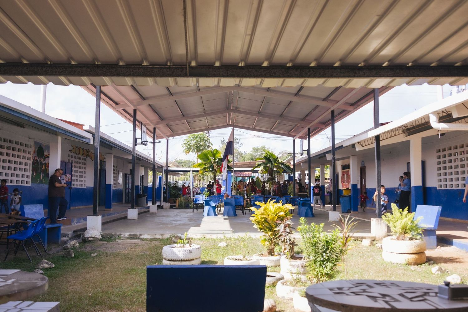 Open-air school courtyard with blue and white buildings, tropical plants, and students gathered under a metal roof.