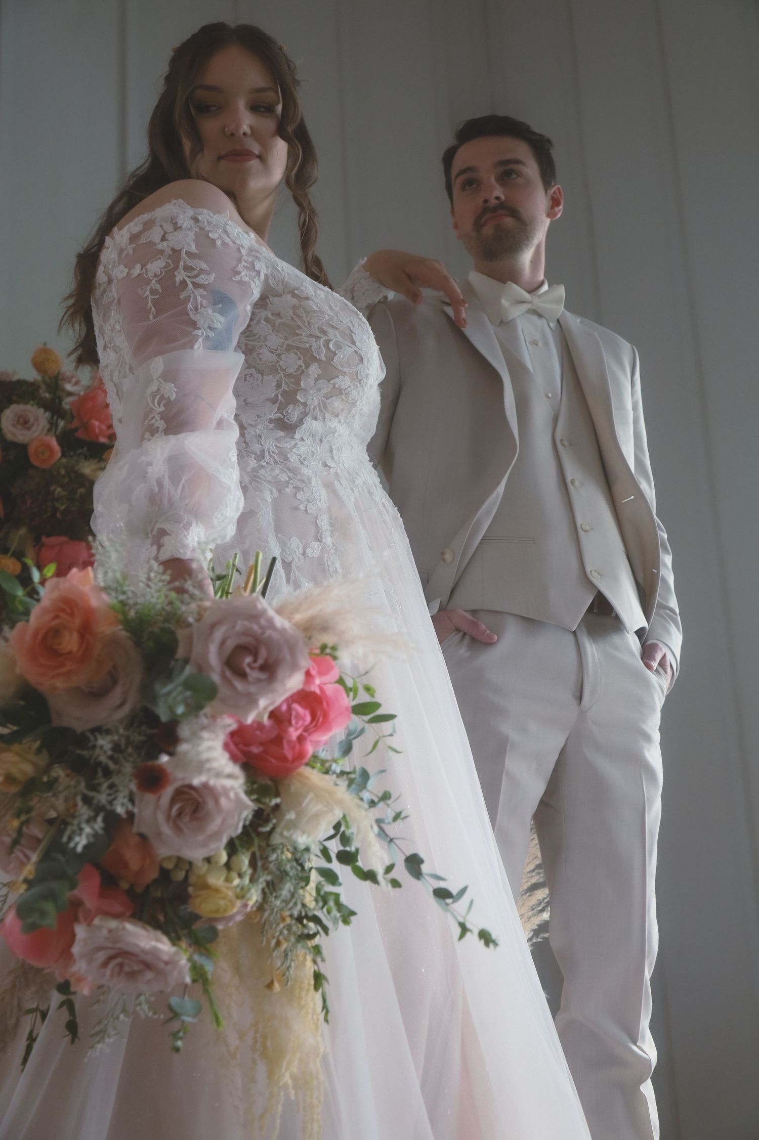 Bride in lace gown stands with groom in white suit holding colorful bouquet with roses in elegant indoor portrait.