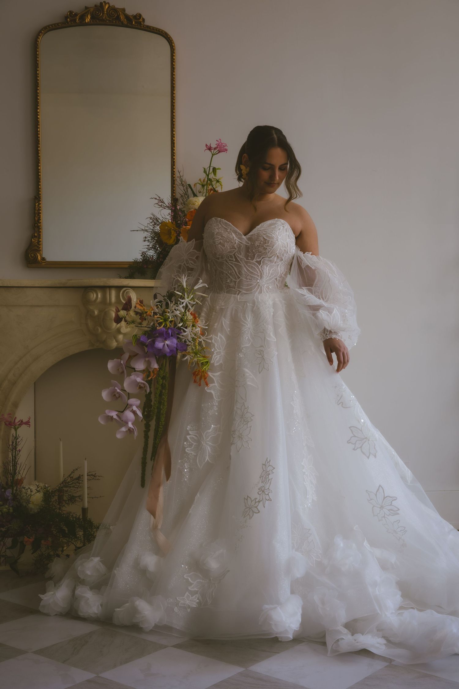 An elegant white wedding gown with floral appliques and off-shoulder sleeves stands by an ornate gold mirror with floral decor.