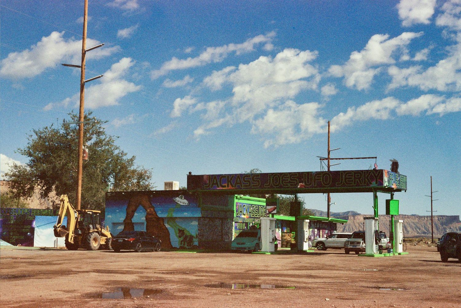 A panoramic desert landscape with small buildings and utility poles under a bright blue sky with white clouds.