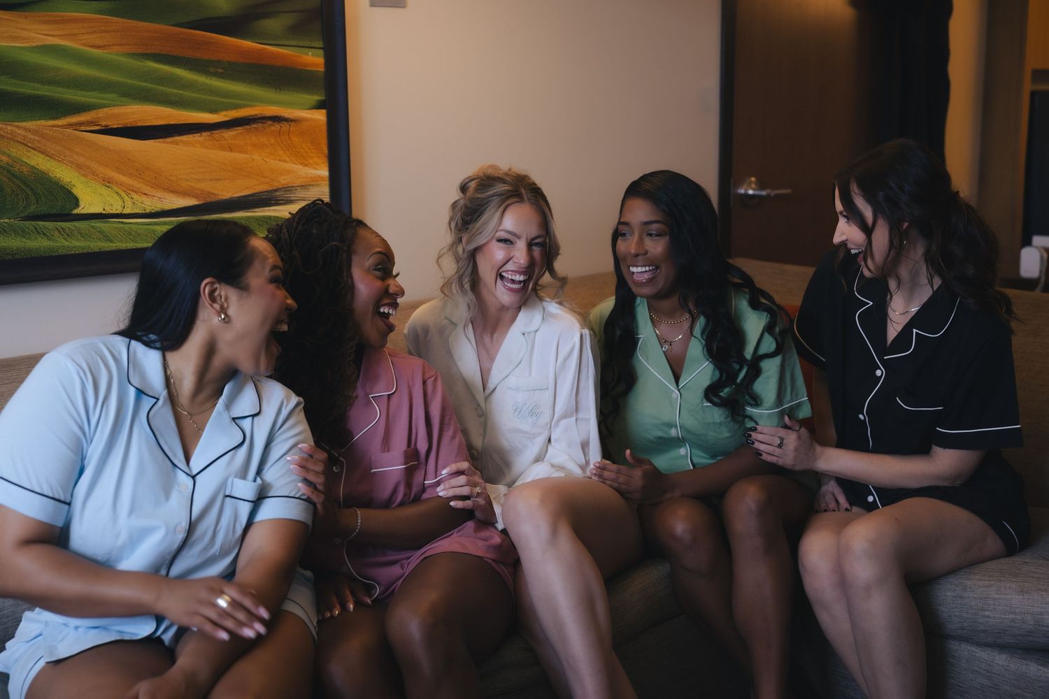 Five women in pajamas laughing together on a couch during a bridal party getting-ready celebration.