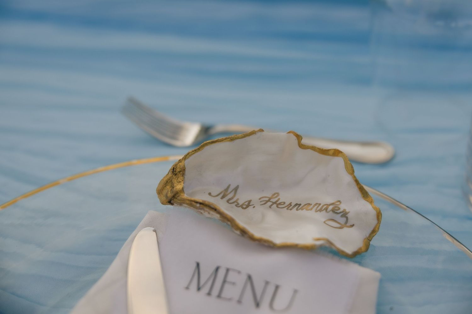 Elegant wedding place setting with a handwritten name on a gold-rimmed oyster shell resting on a menu card.
