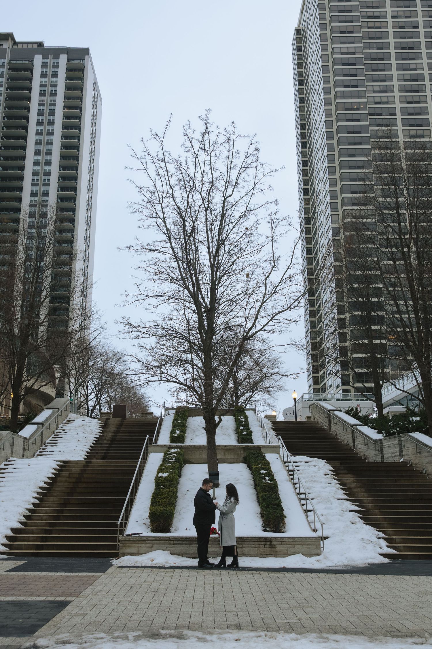 Two figures stand at the base of a snowy staircase between tall apartment buildings in an urban winter landscape.