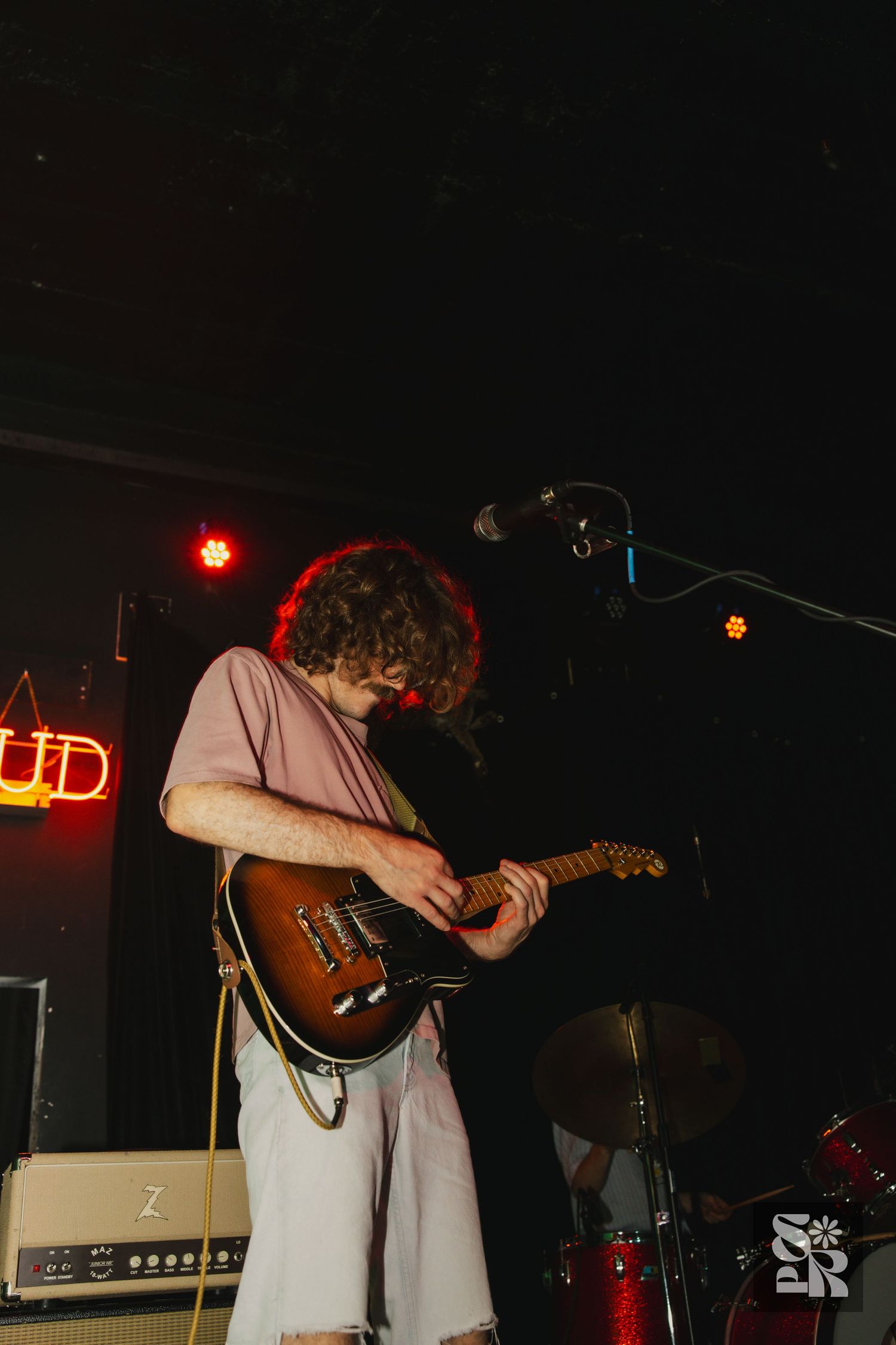 Guitarist performing on stage in a low-lit venue wearing a light-colored shirt while leaning over their instrument.