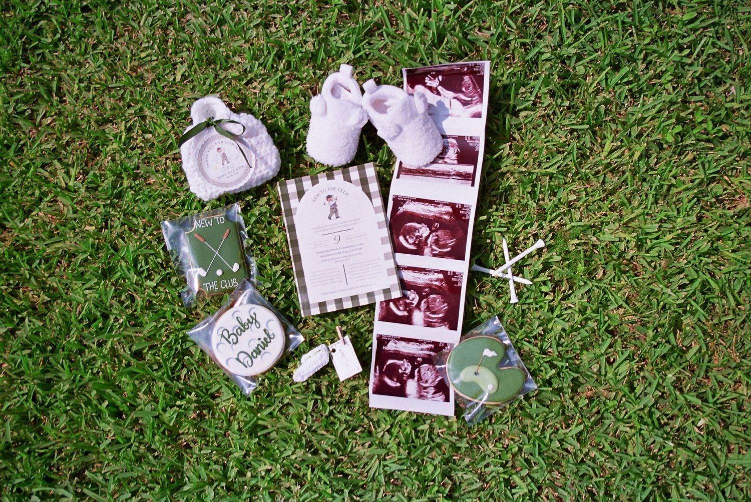 Baby shower flat lay with ultrasound photos, tiny shoes, cookies, and gifts arranged on green grass.