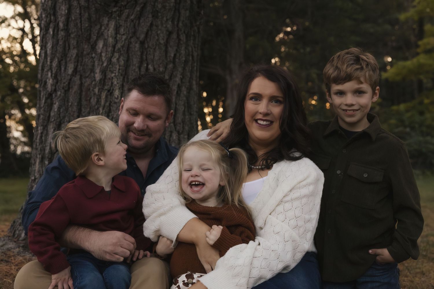 A happy family of five poses outdoors near a large tree, smiling together in a warm autumn setting.