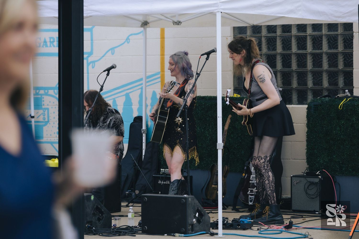 Three female musicians performing on an outdoor stage under a white tent, playing guitars at a live music event.