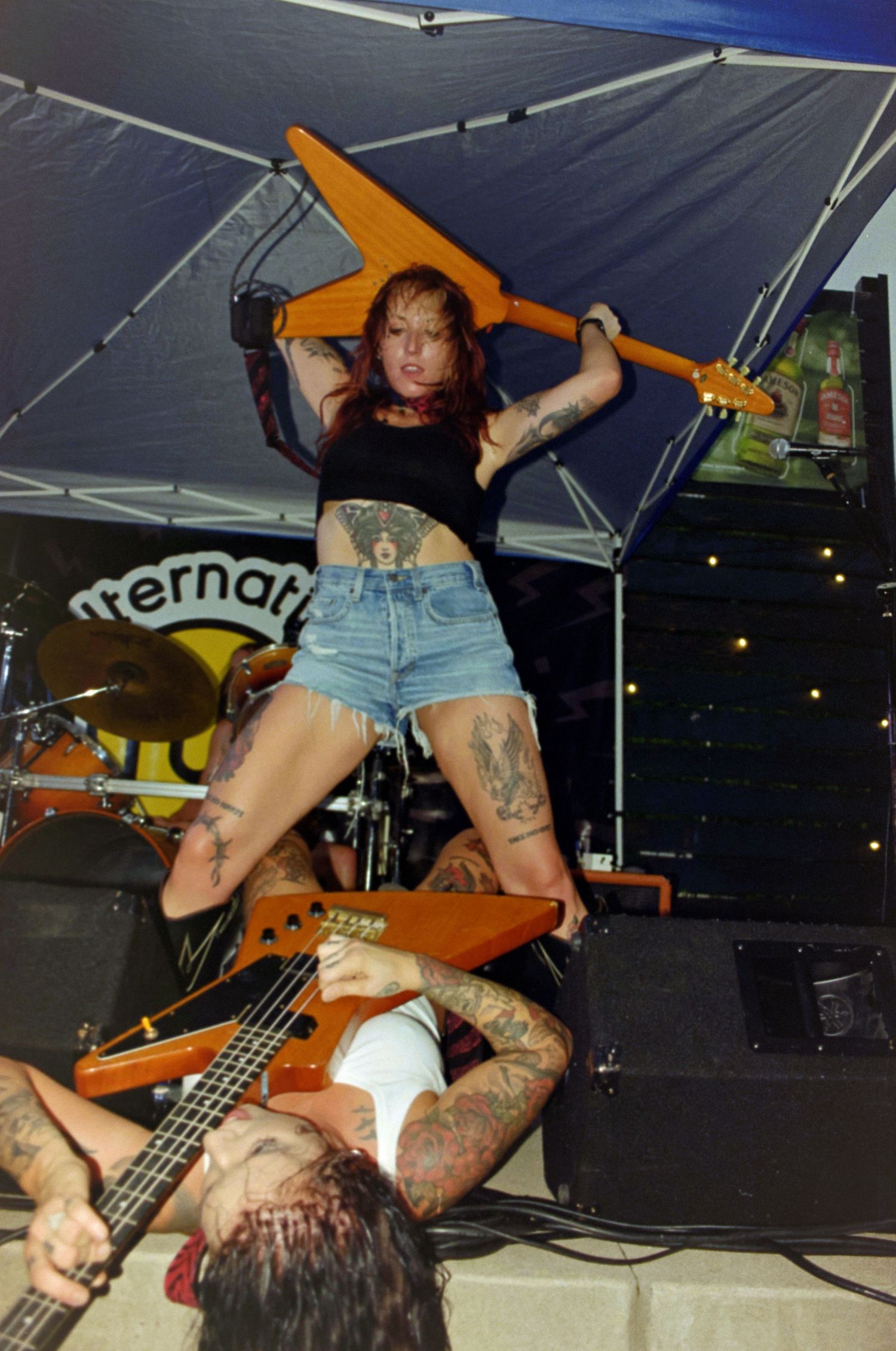 Tattooed female punk rock guitarist performing on stage, raising orange guitar overhead while standing over another musician.