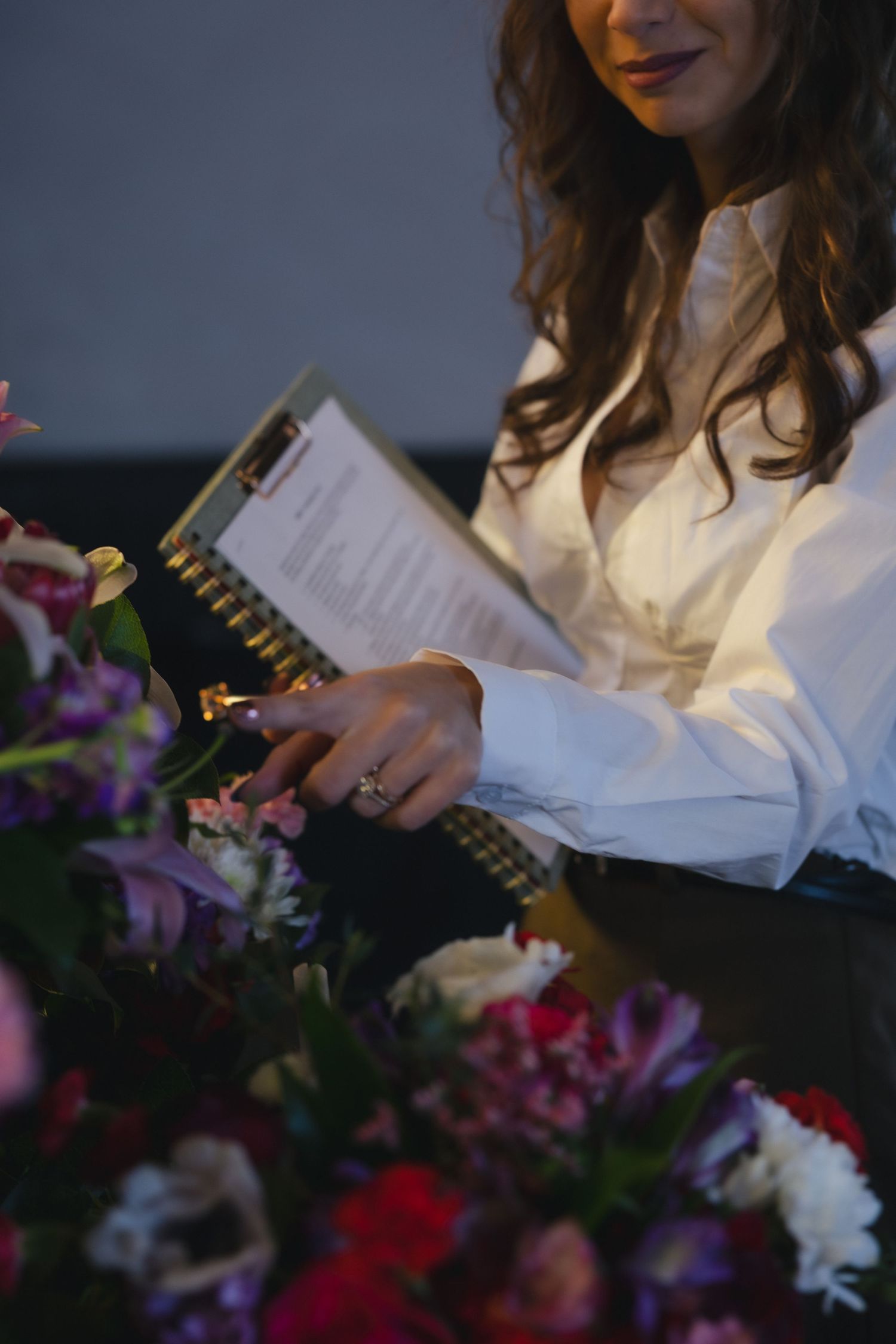 Woman in white blouse arranging colorful flowers while holding a clipboard at an elegant floral event.