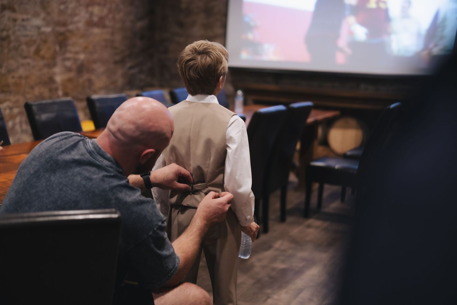 Man helping young boy adjust his vest and tie in a rustic venue before a formal event.