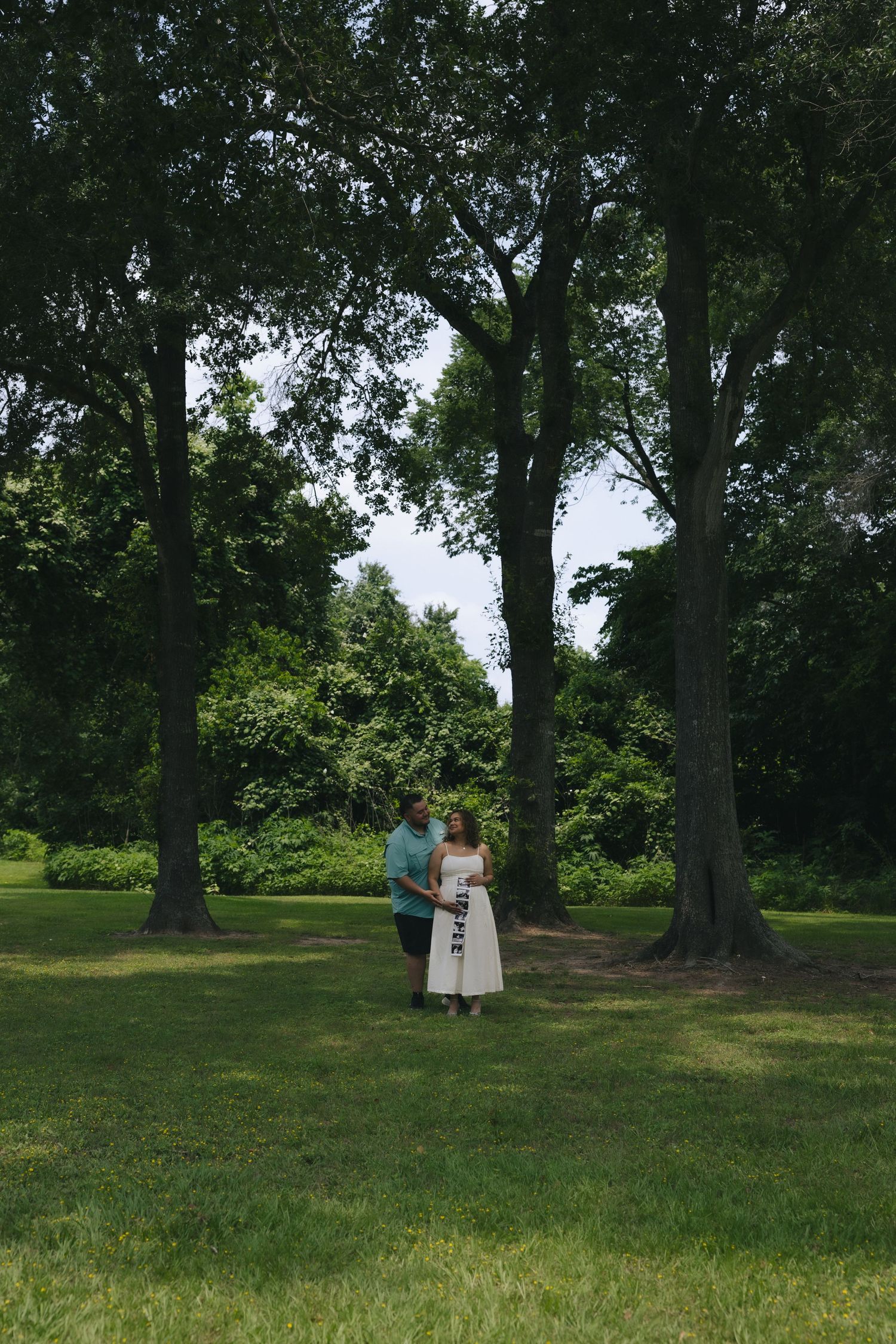 Couple standing together in a lush green park surrounded by tall trees on a summer day.