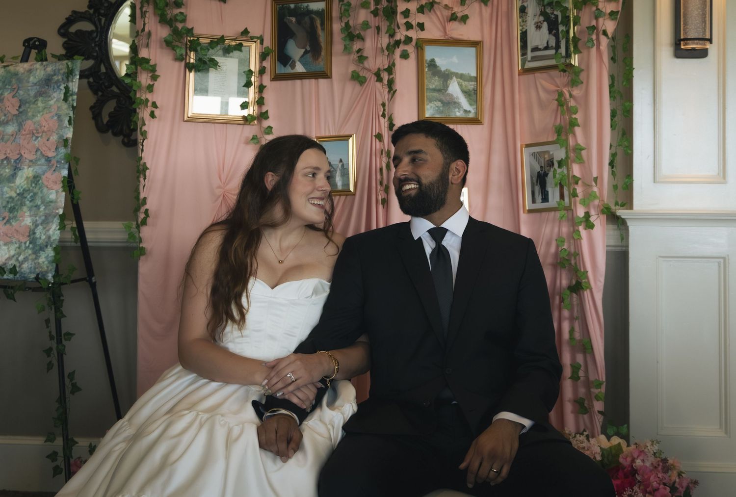 A bride and groom sit together smiling, dressed in wedding attire, against a pink floral backdrop.