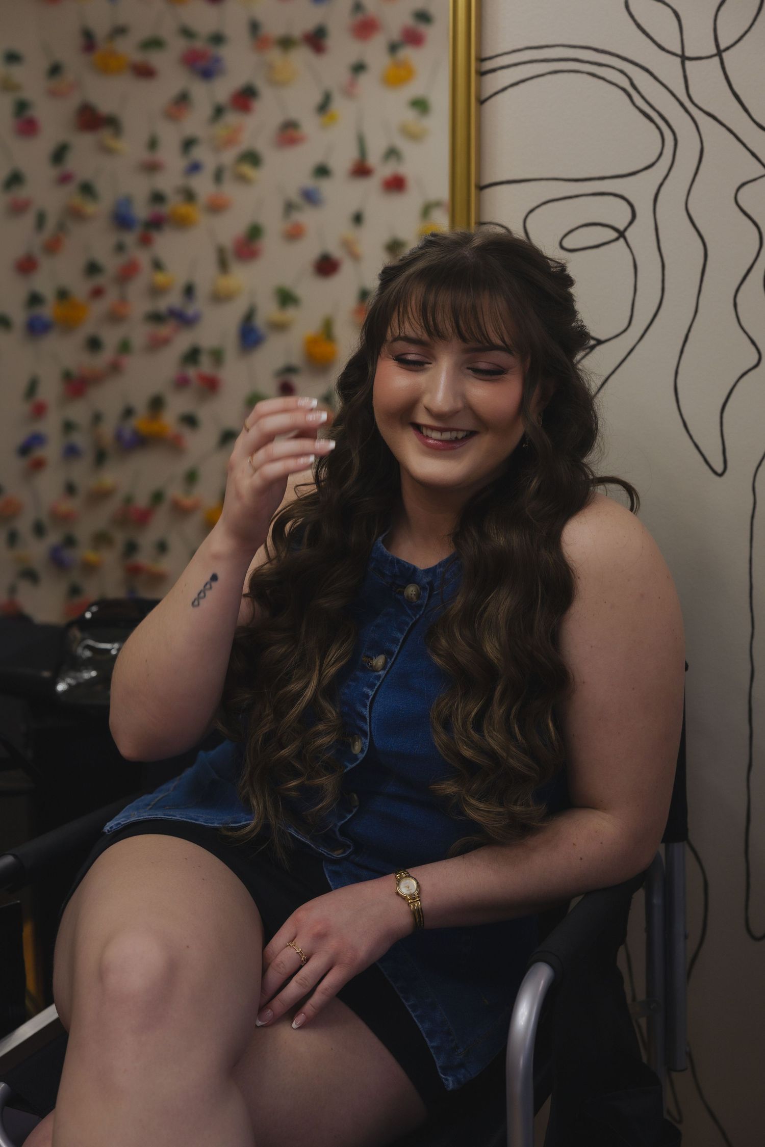 Smiling young woman with long wavy hair sitting in a salon chair wearing a denim top and shorts.